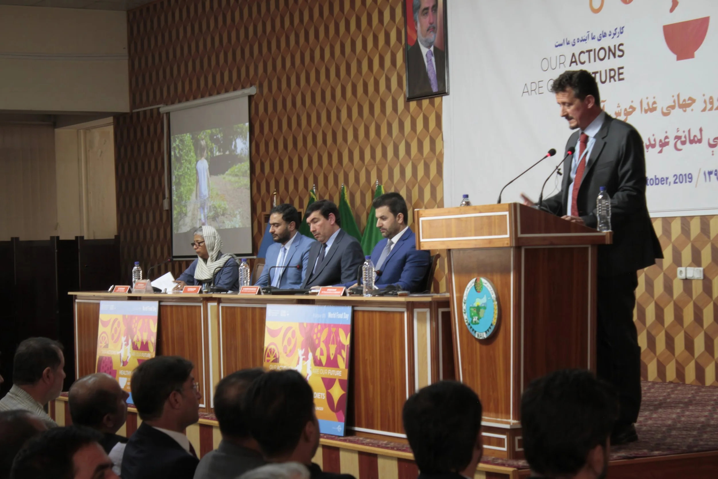 A man in a suit is speaking at a podium during a formal event, with four other individuals seated at a long table behind him. The background includes a large white banner with text and graphics, a framed photo of a man on the wall, and a projection s