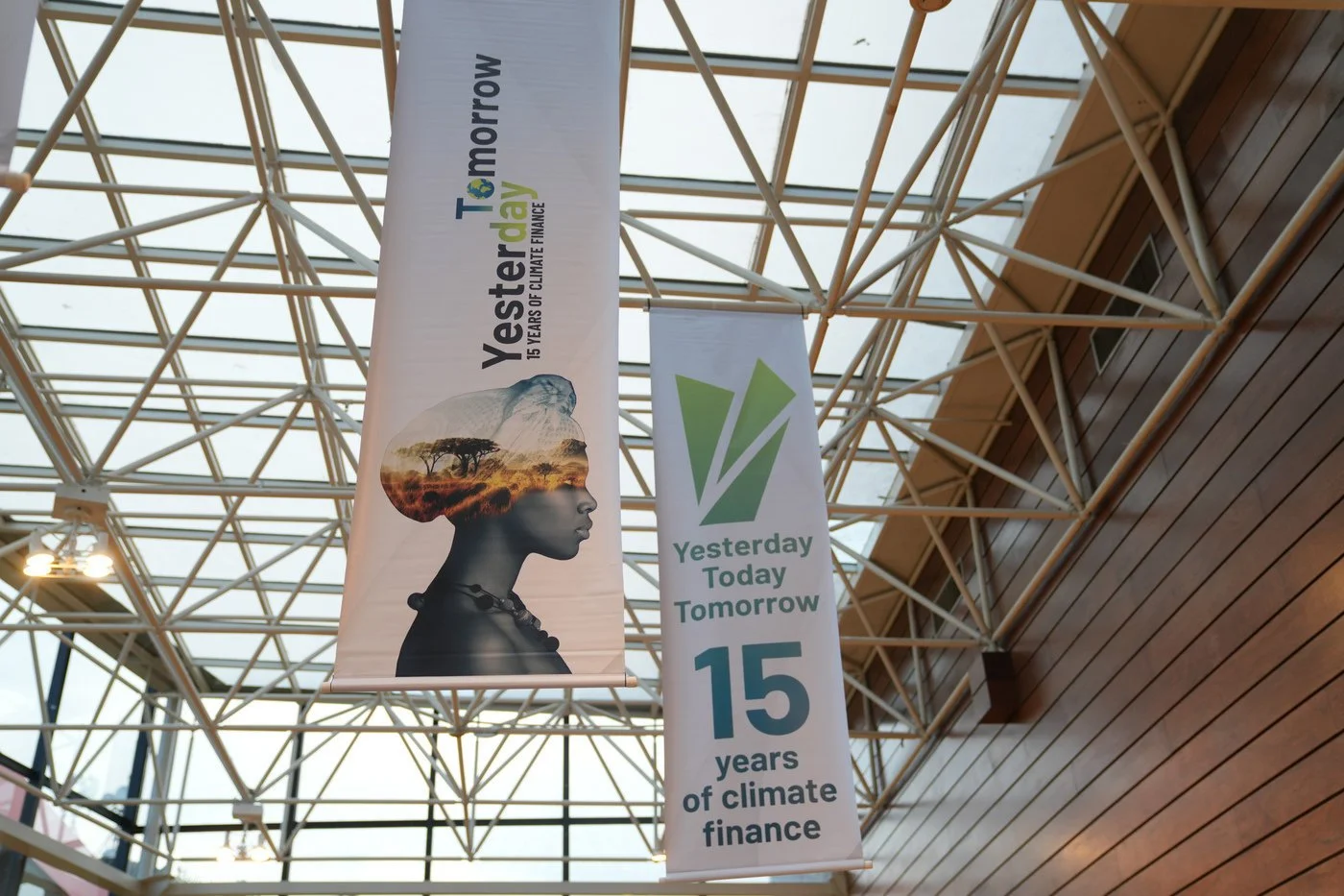 Two vertical banners hanging inside a building with a glass ceiling and wooden wall. The left banner features a silhouette of a woman with a landscape of trees and the earth's surface inside her head, with text about climate finance. The right banner