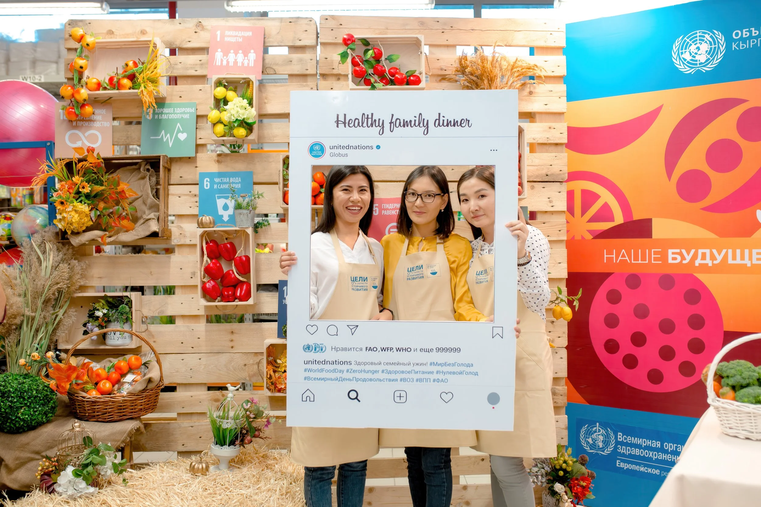 Three women are holding a large fake social media frame at a health fair, with a backdrop of wooden crates filled with artificial fruits and vegetables, and banners promoting healthy eating and the United Nations.