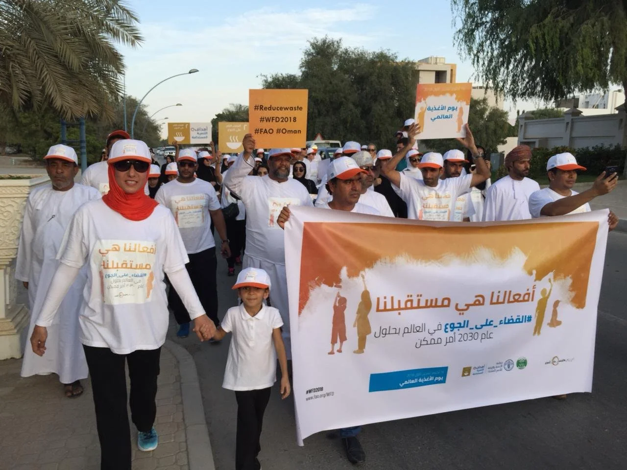 Group of people participating in an outdoor walk for environmental awareness, holding a large banner with Arabic text and smaller signs promoting waste reduction and global environmental goals.