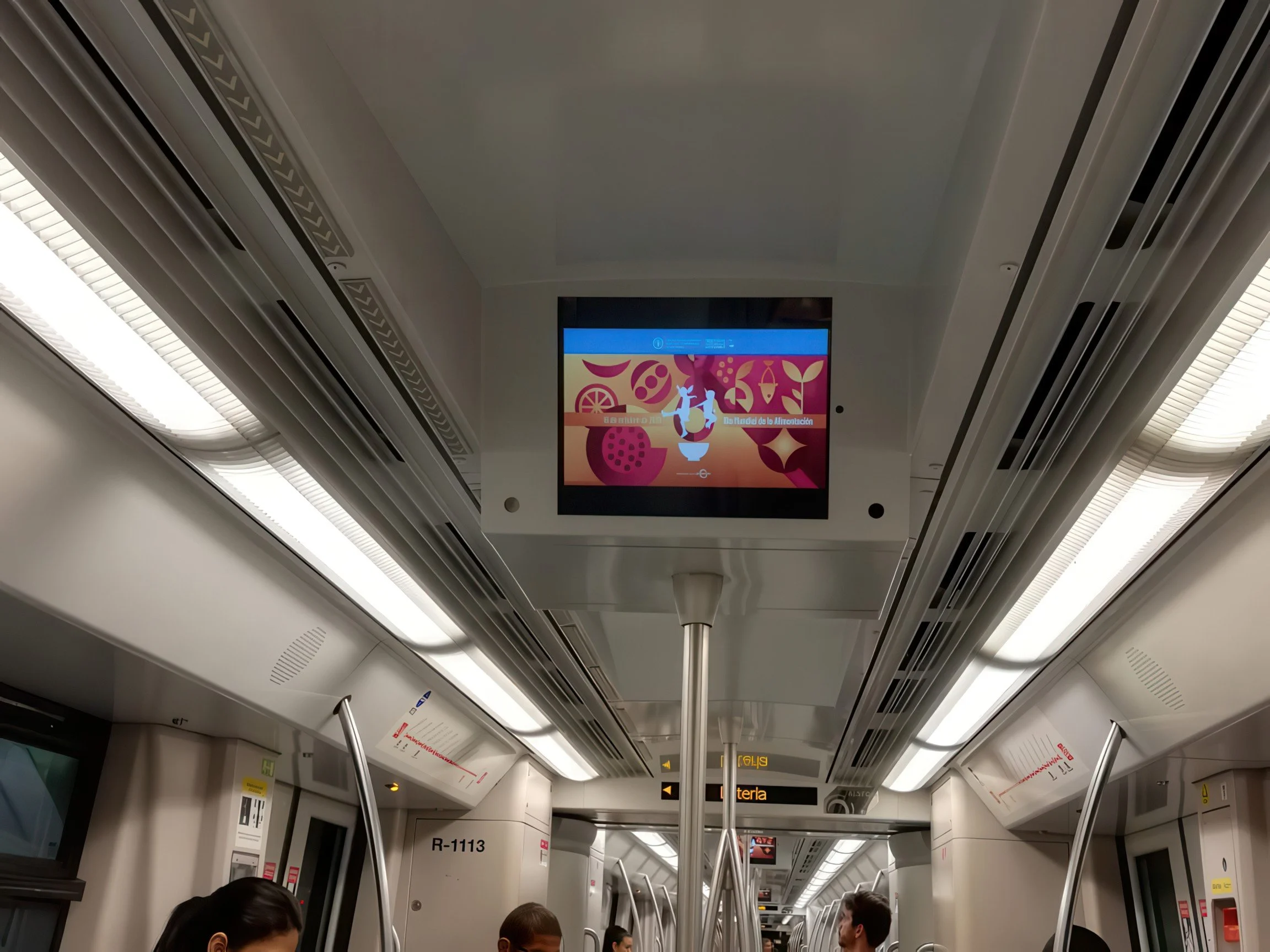 The interior of an aircraft cabin with overhead compartments, digital screens, and seated passengers.