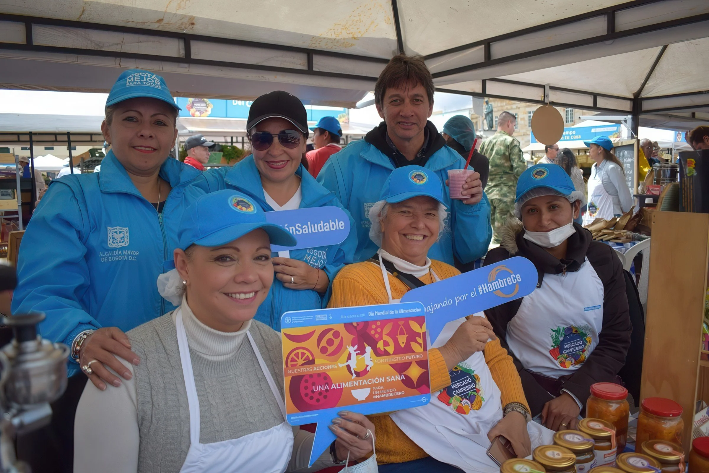 Group of six smiling people, including women and a man, at an outdoor event or market, wearing blue jackets and some with blue hats, holding signs that promote healthy eating and nutrition, with tables of jars in front of them.