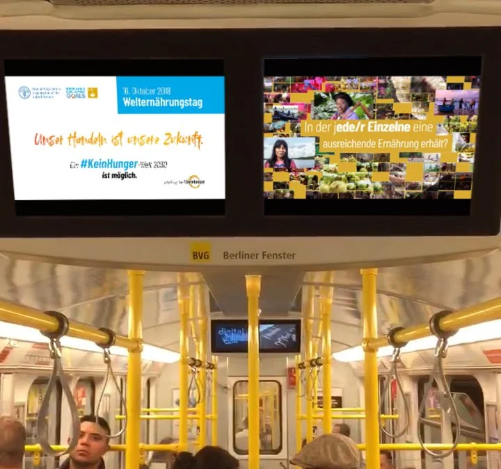 Inside a subway train, showing digital screens with advertisements and information, with passengers seated and standing, yellow handrails, and a curved ceiling.