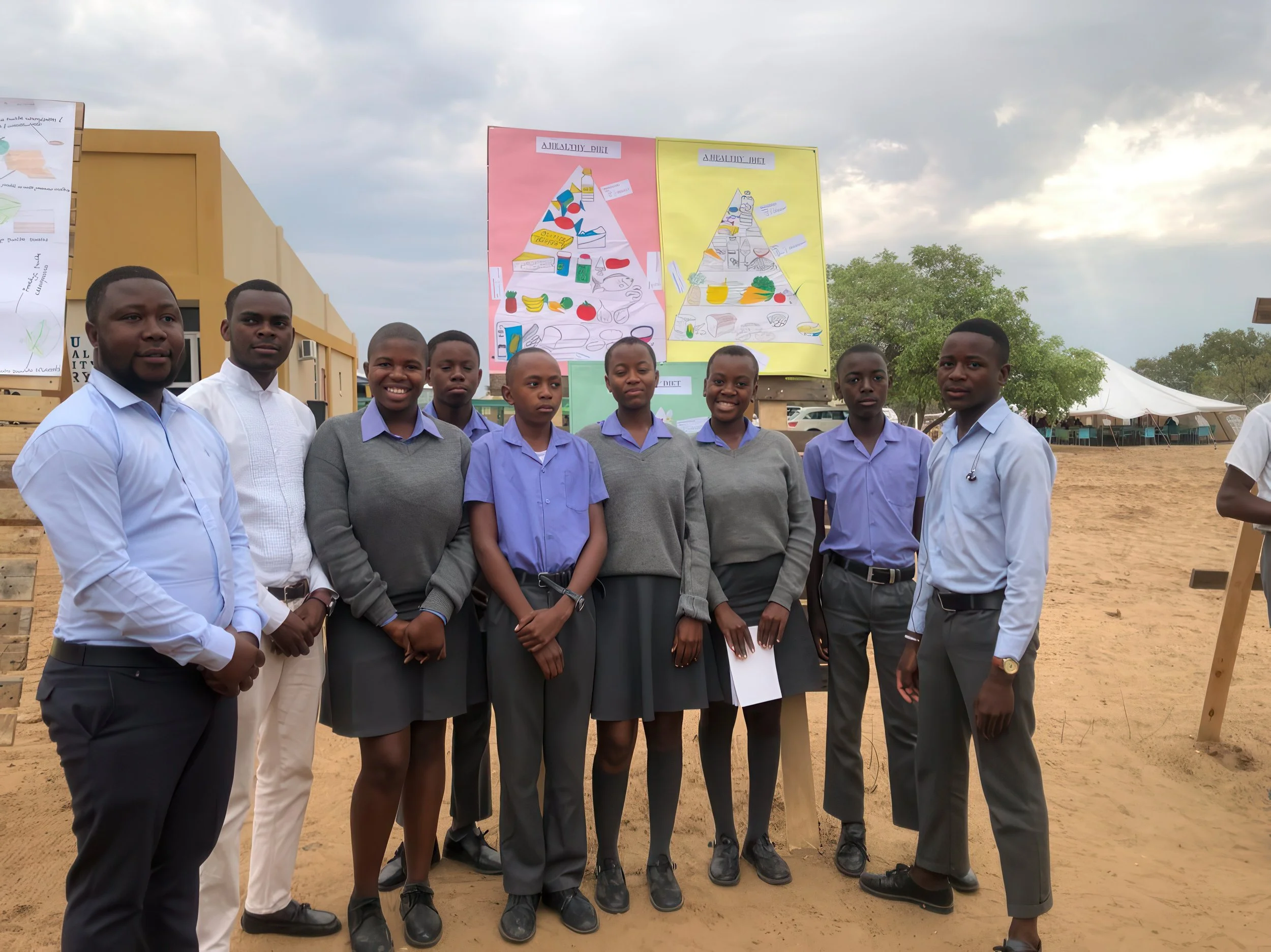 A group of nine young students, six boys and three girls, standing outside on a dirt ground. They are dressed in school uniforms with some in light shirts and others in darker, grayish or purple shirts. Behind them are two large colorful posters on d