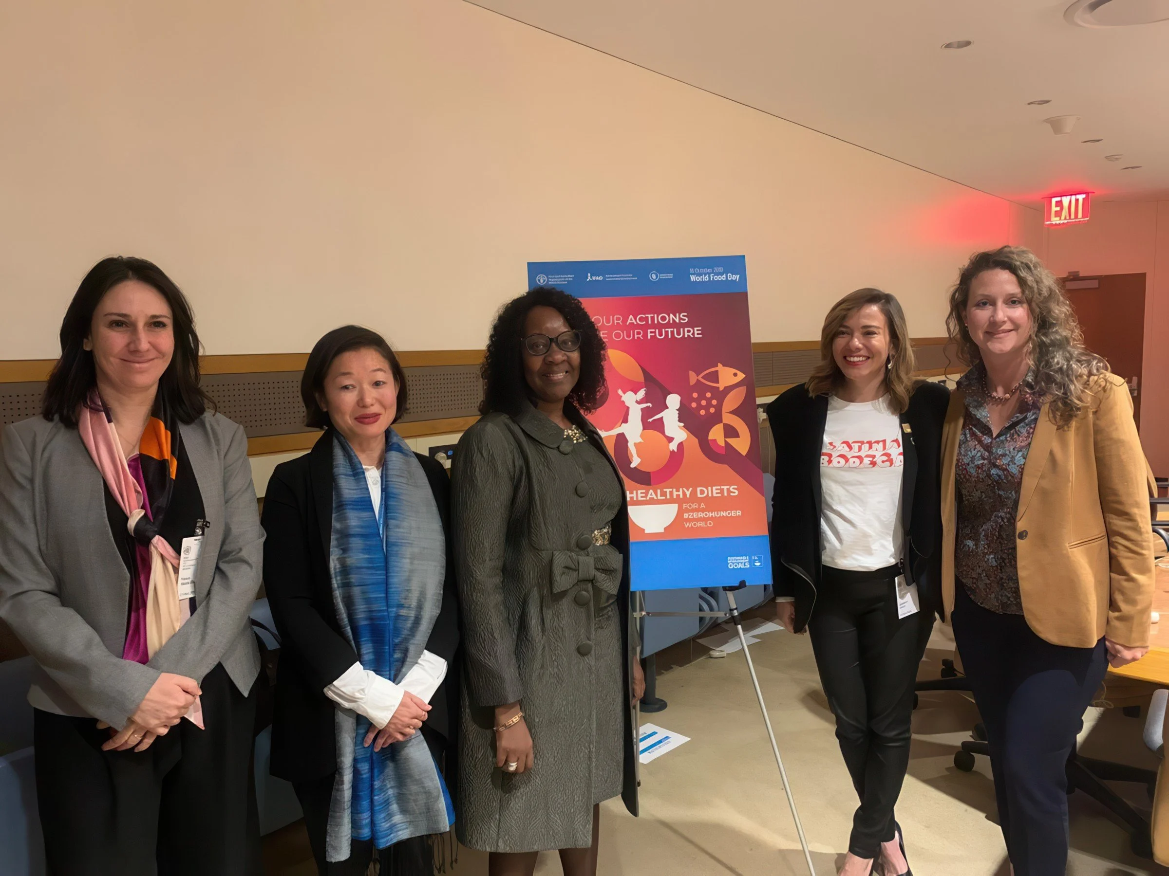 A group of six women standing side by side in front of a poster at a conference or event. The poster mentions World Food Day and healthy diets. The women are smiling and dressed in business attire.