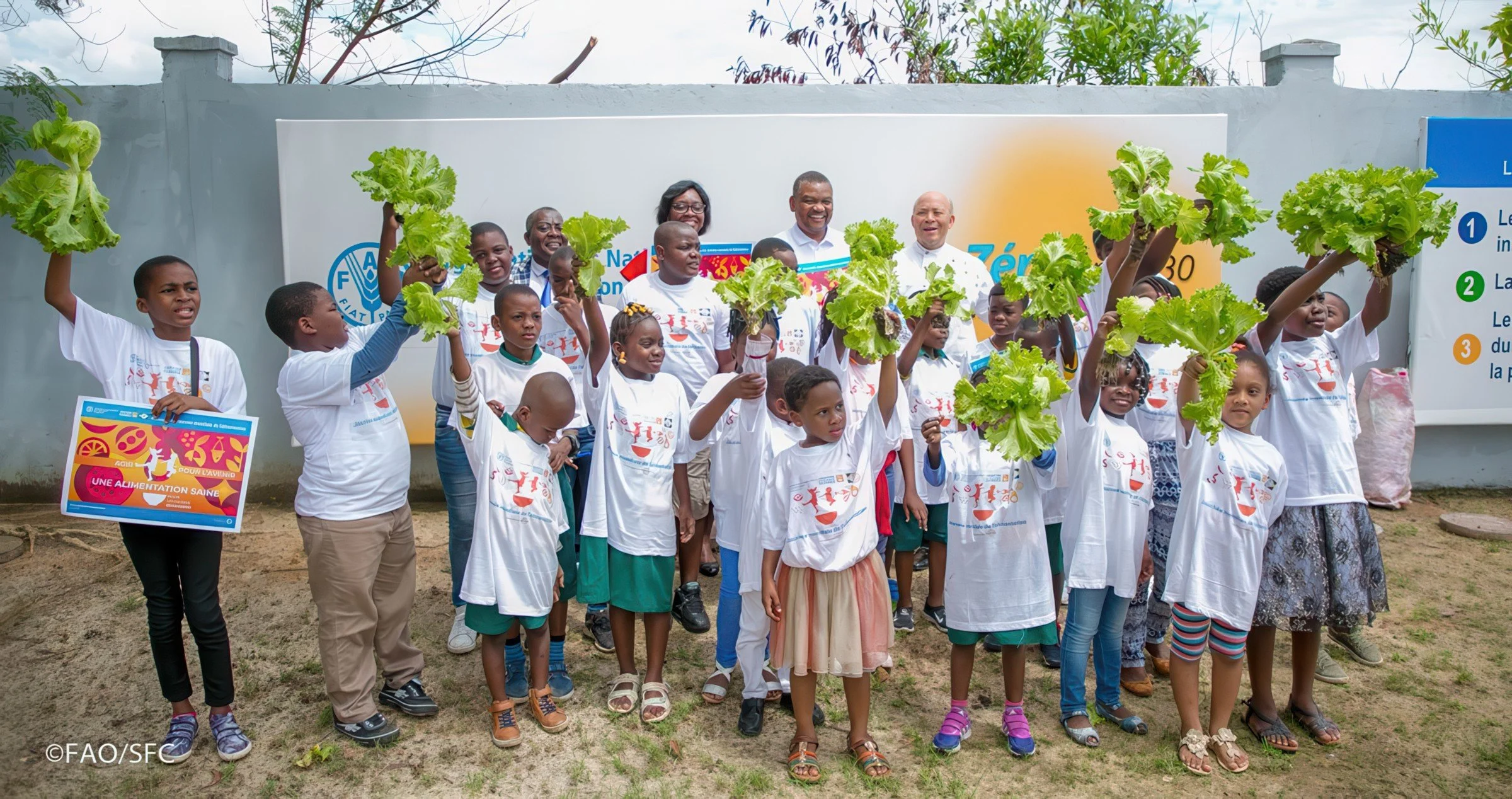 Group of children and adults outdoors holding up lettuce heads during a community event or celebration. Some children hold signs related to healthy eating. There are banners and a white wall in the background.