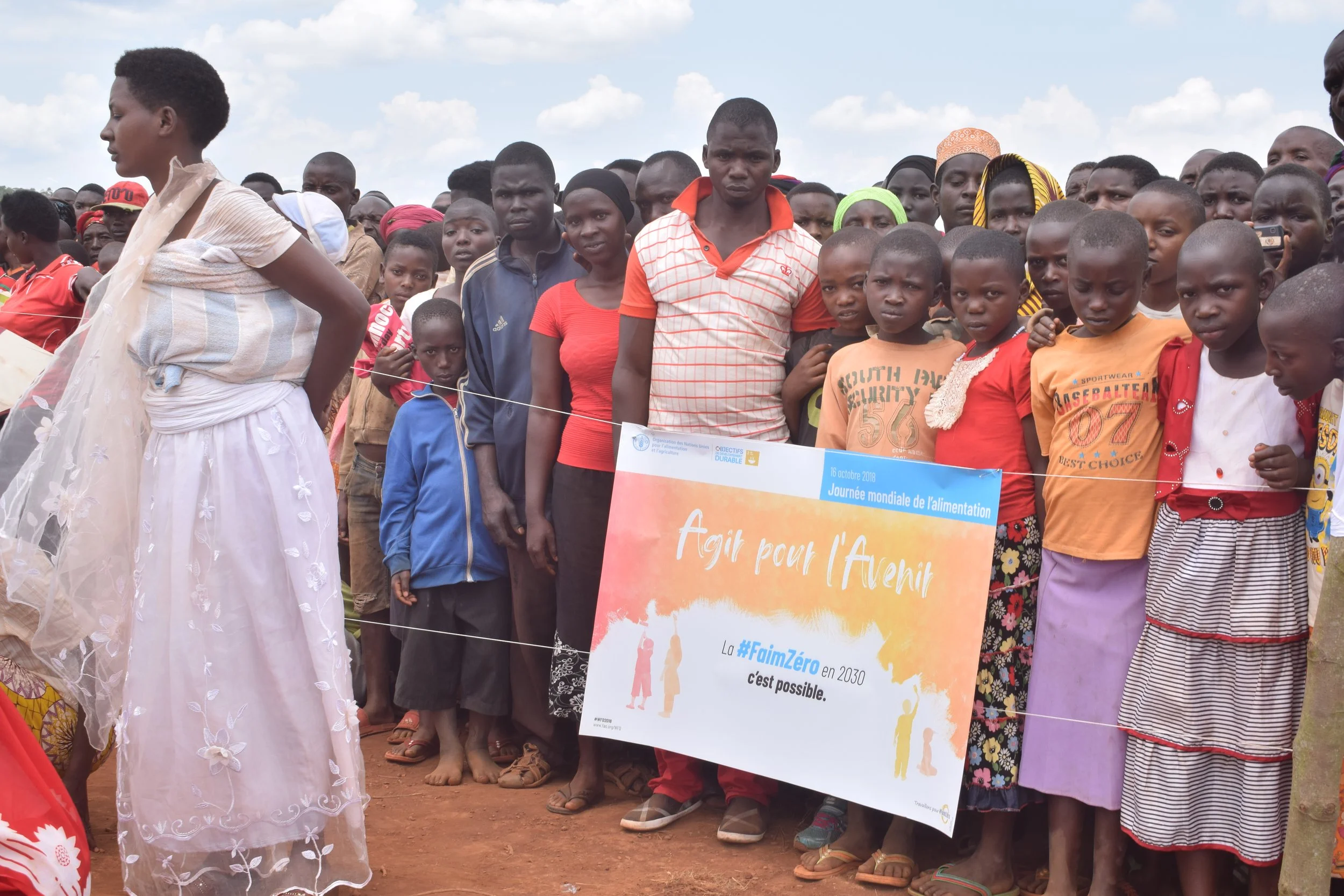 A group of people, including children and adults, gather outdoors on a dirt ground against a cloudy sky. Some of them are holding a banner with French text about a global food day and futuristic food security goals, with a UNICEF logo and date of Oct