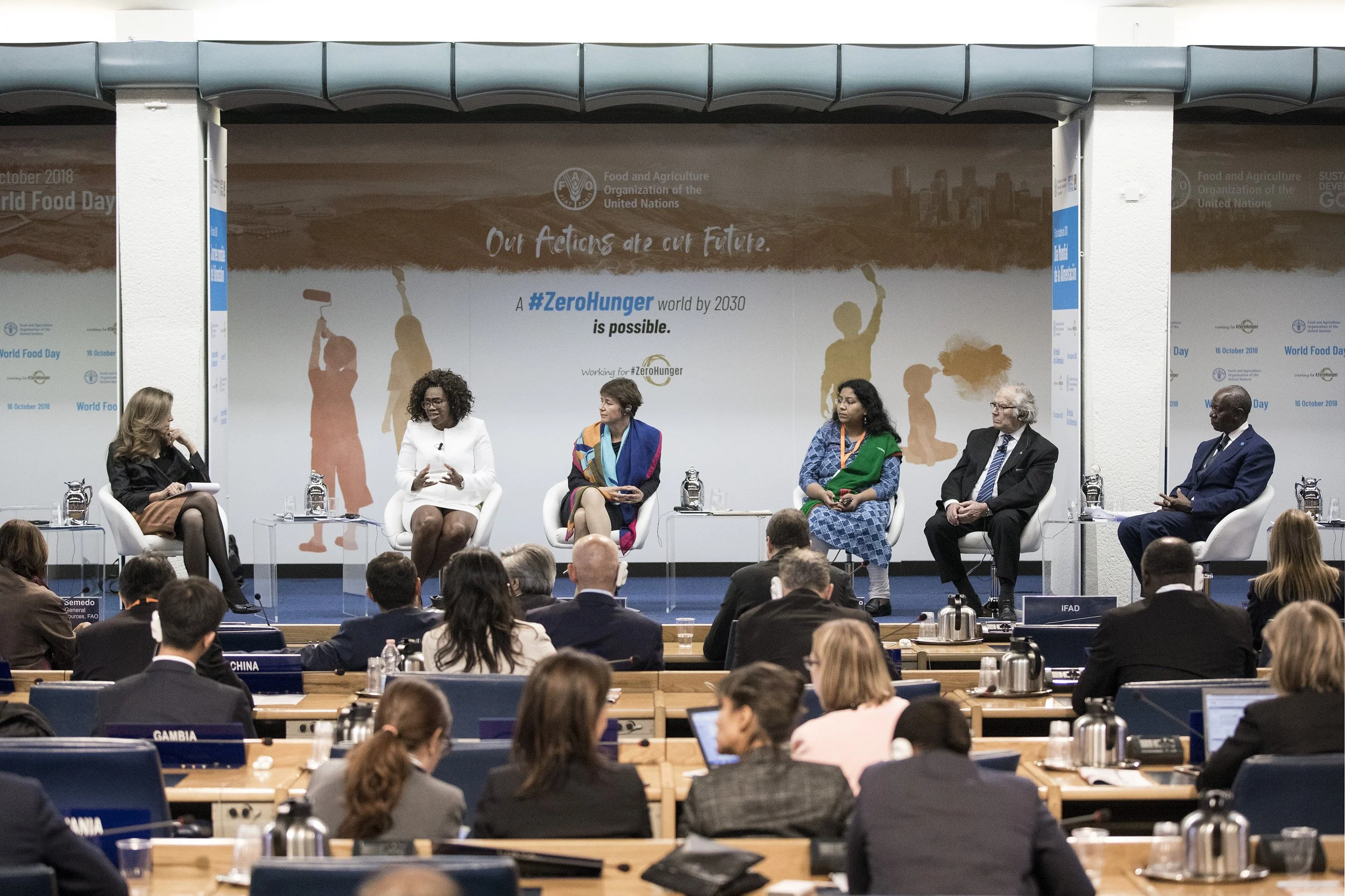 Panel discussion at a conference with five women seated on stage, with the words 'Our Actions are our Future' and '#ZeroHunger' displayed on a large screen behind them. Audience members are visible in the foreground.