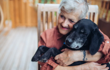 An elderly woman hugging a black dog inside a cozy wooden room.