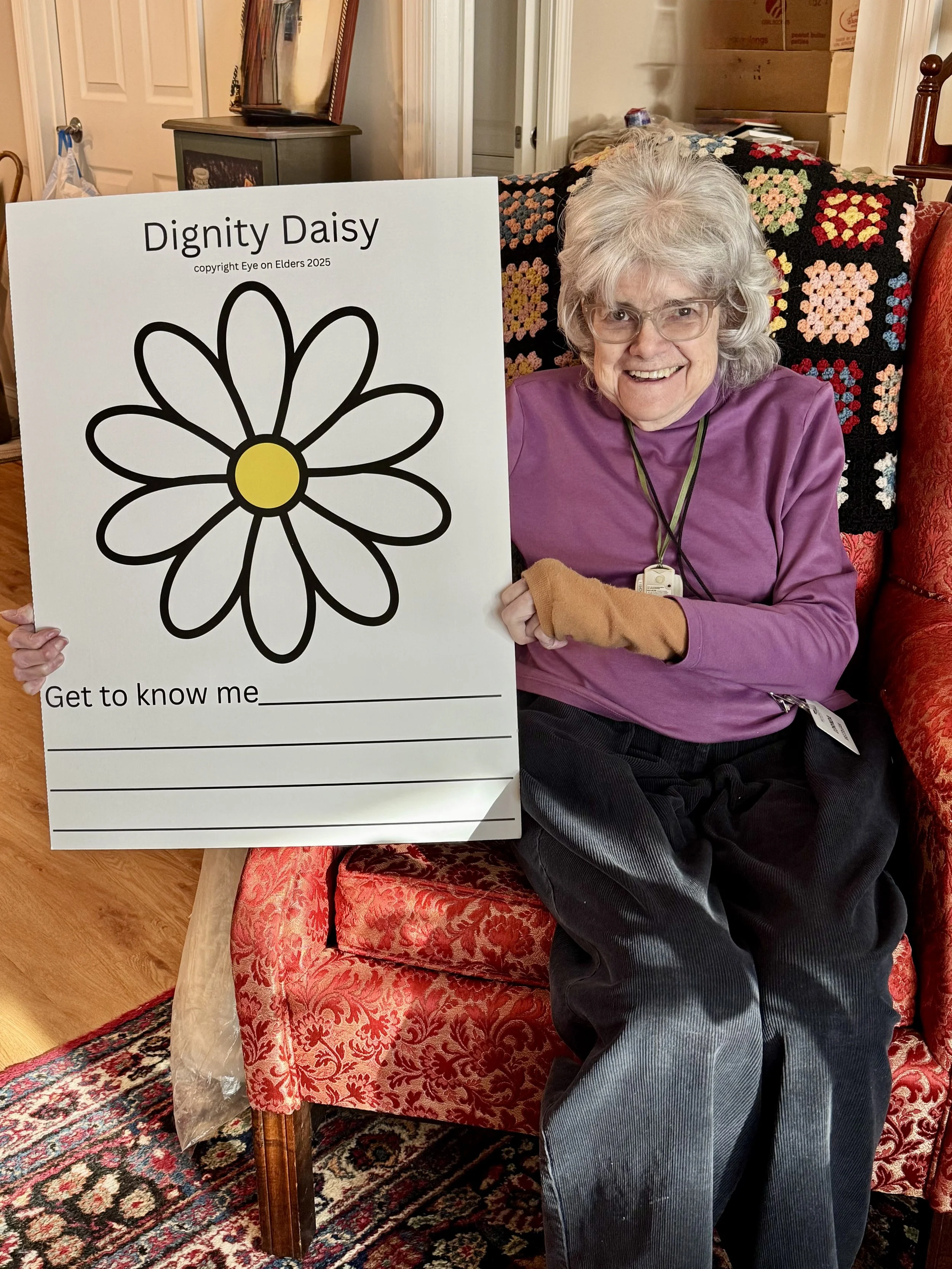 A smiling elderly woman with gray hair and glasses sitting on a red patterned armchair and holding a sign that says 'Dignity Daisy' with a daisy flower illustration and the words 'Get to know me'