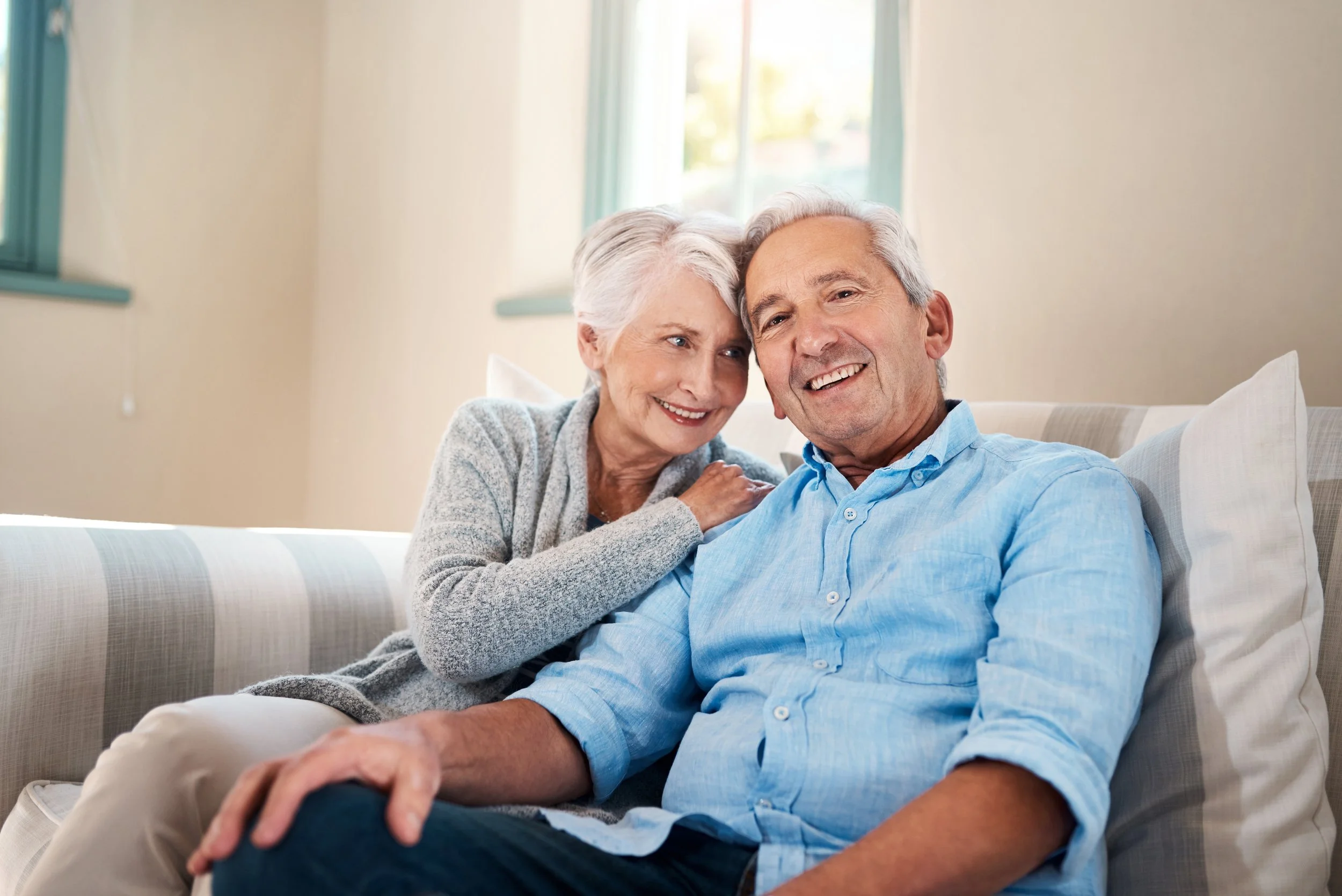 An elderly woman and man sitting close together on a couch, smiling and enjoying each other's company in a well-lit room with a window in the background.
