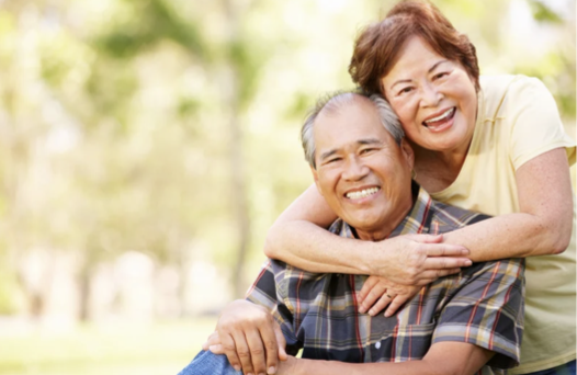 A smiling older man and woman enjoying a hug outdoors in a park.