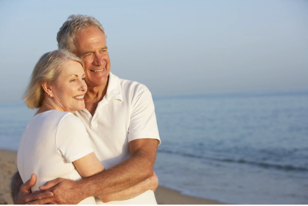 An elderly couple smiling and hugging on a beach with the ocean in the background.