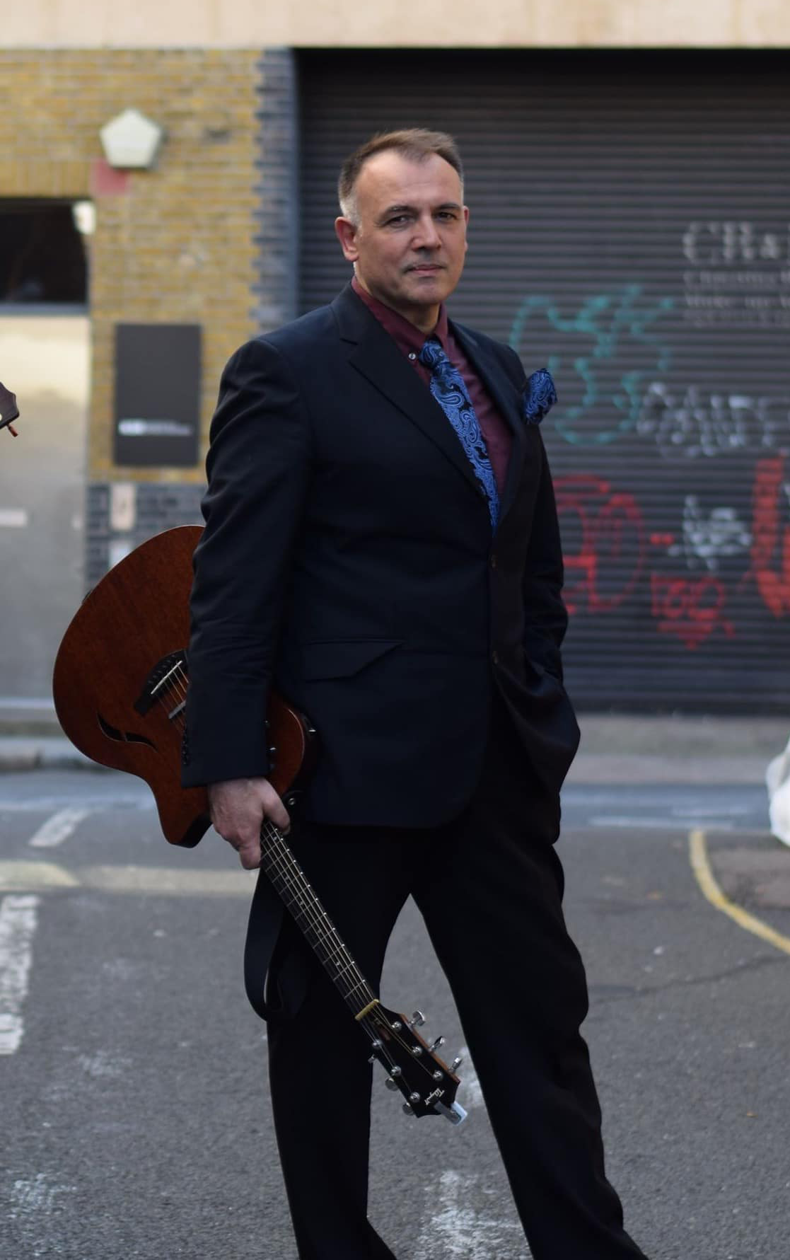 A man in a black suit holding an acoustic guitar, standing outdoors with a brick building and graffiti in the background.