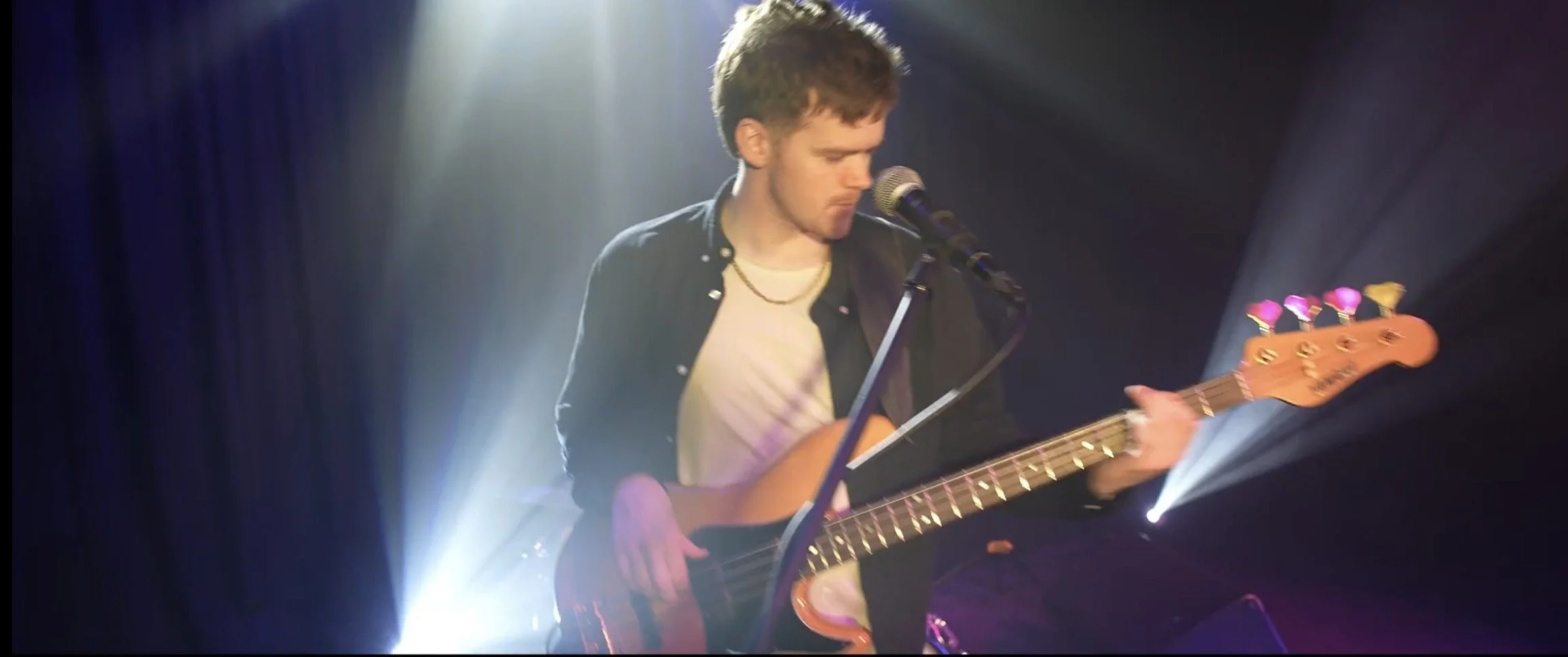 A young man with brown hair, wearing a black jacket and white shirt, playing an electric guitar on stage with bright lights and a dark background.