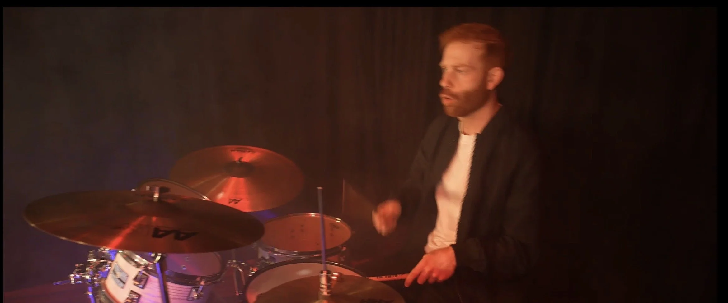 A man with a beard playing drums on stage, illuminated by red and blue lighting, with a dark background.