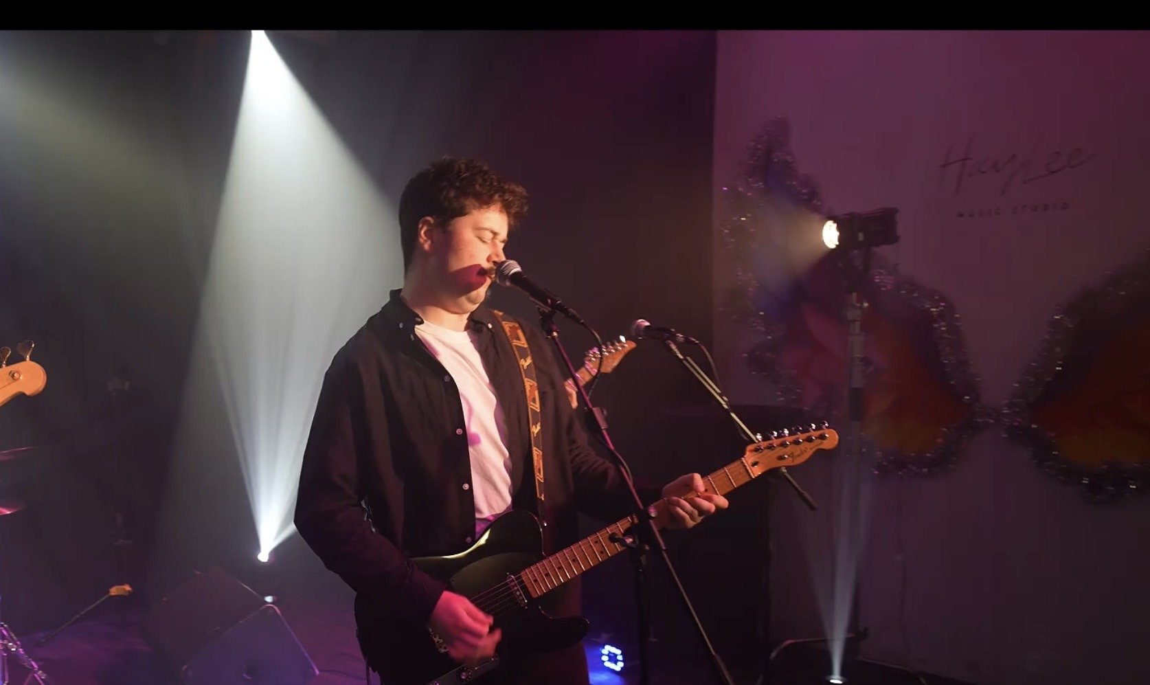 A young male musician playing an electric guitar and singing into a microphone on stage during a performance, with stage lighting creating a spotlight effect.