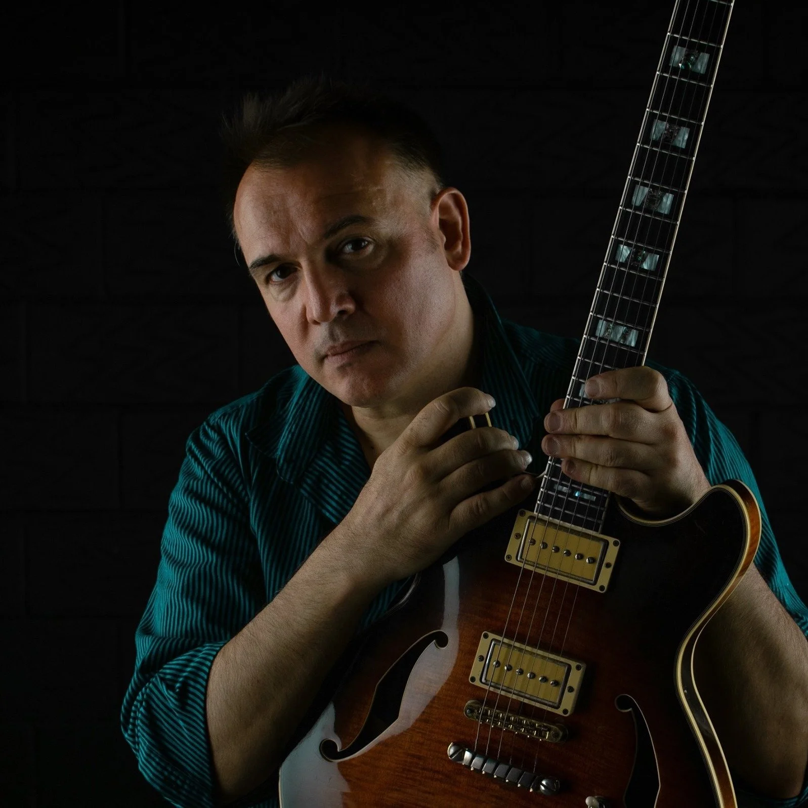 A man with dark, short hair and a serious expression is holding an electric guitar while looking at the camera. He is wearing a teal striped shirt and is in a dark room.
