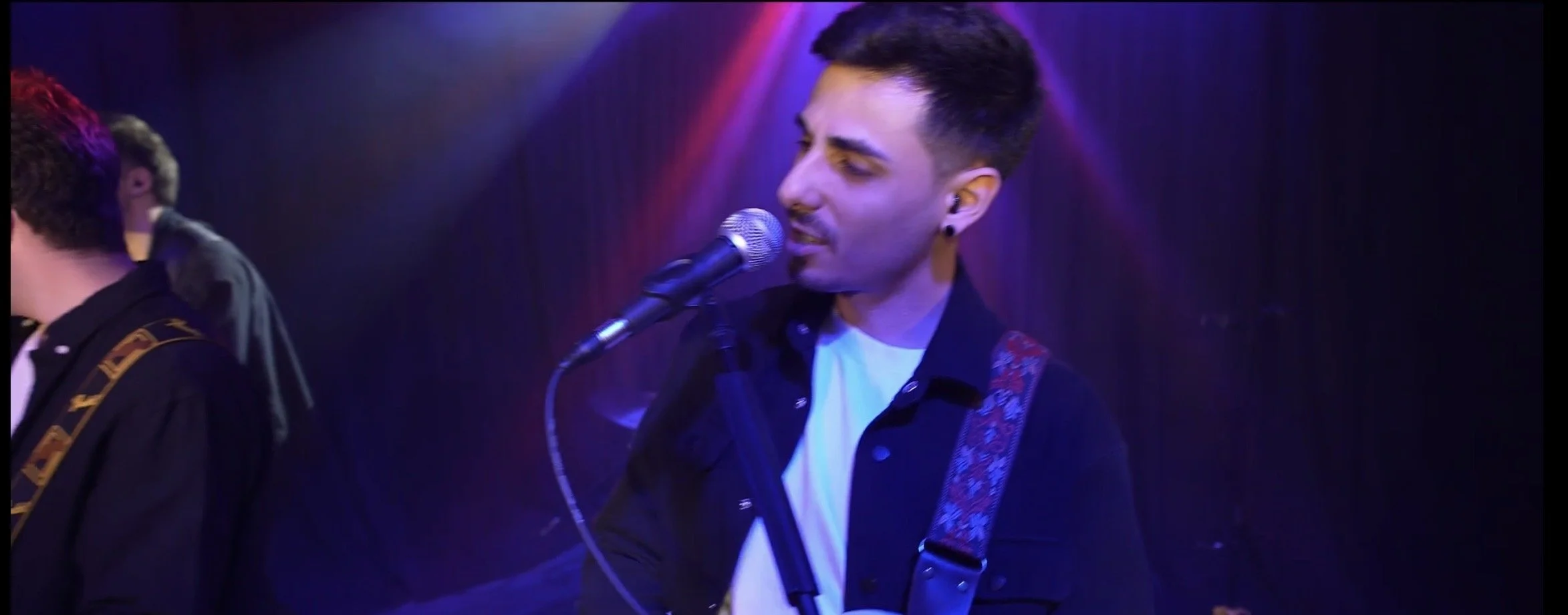 A young man singing into a microphone on stage with colorful lighting, wearing a black jacket, white shirt, and a patterned guitar strap.