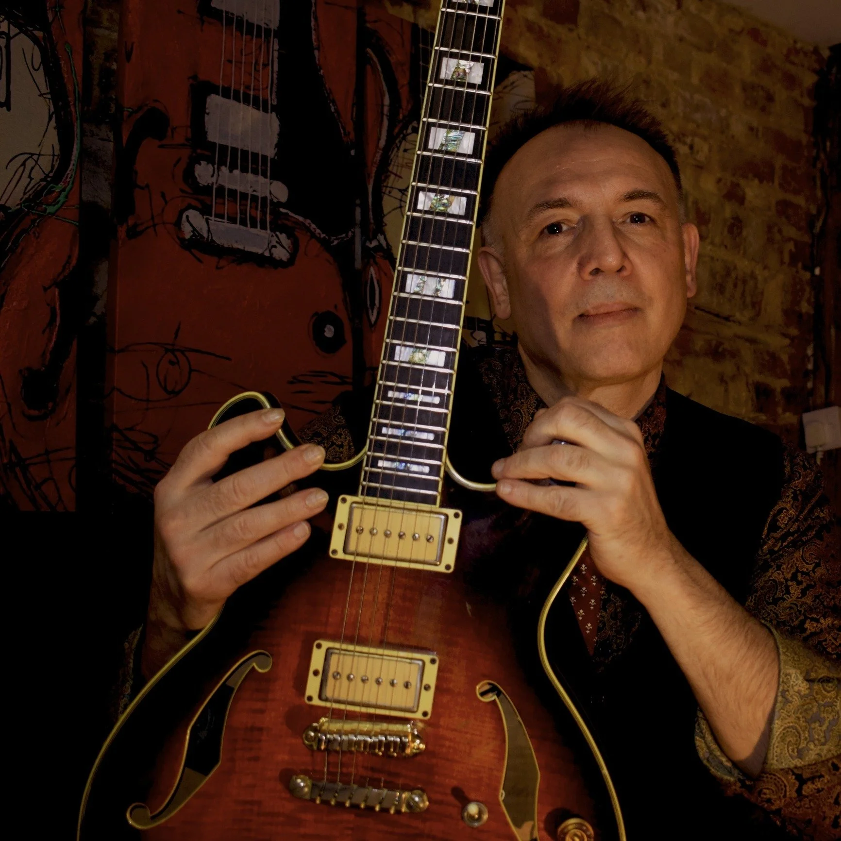 A middle-aged man with short dark hair and a light complexion holding a guitar, with a brick wall and a painted artwork of a dog's face in the background.