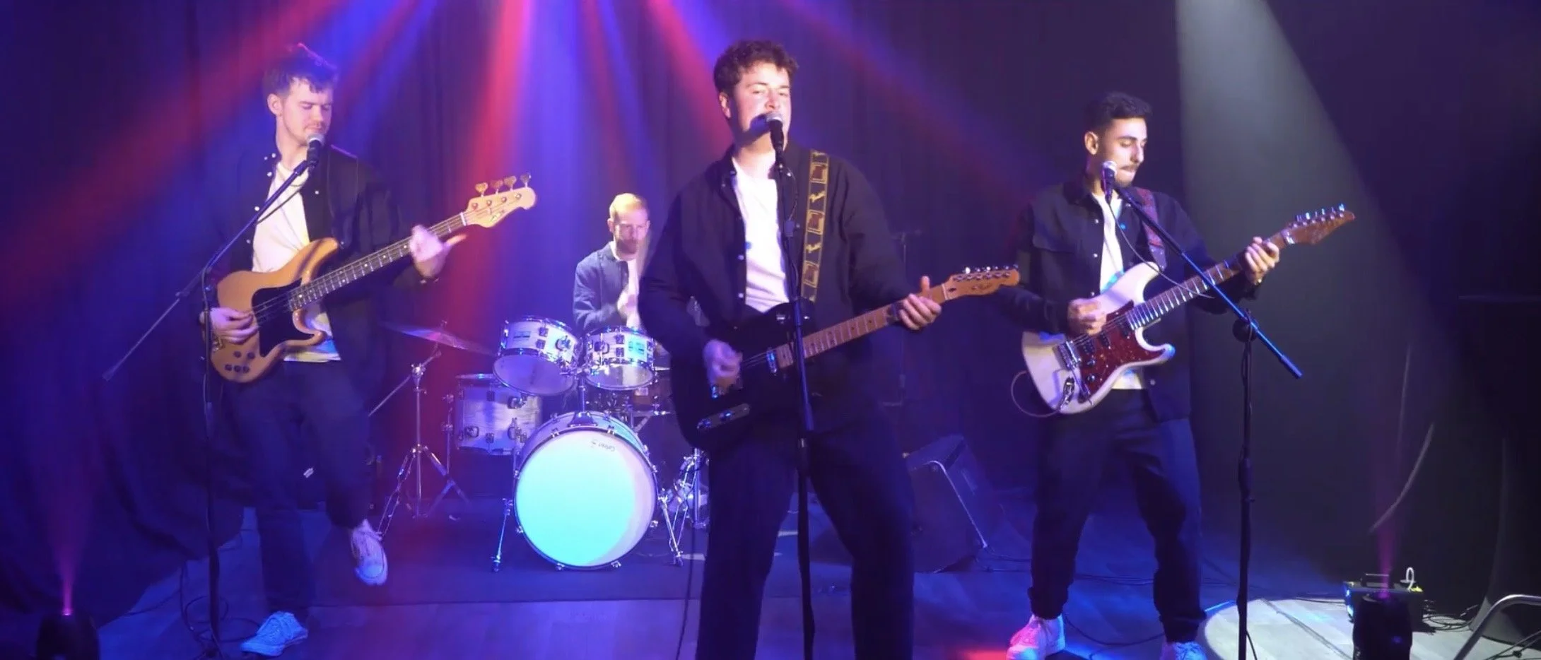 A four-member band performing on stage with colorful lighting. Two members are playing electric guitars, one on drums, and one singing into a microphone.