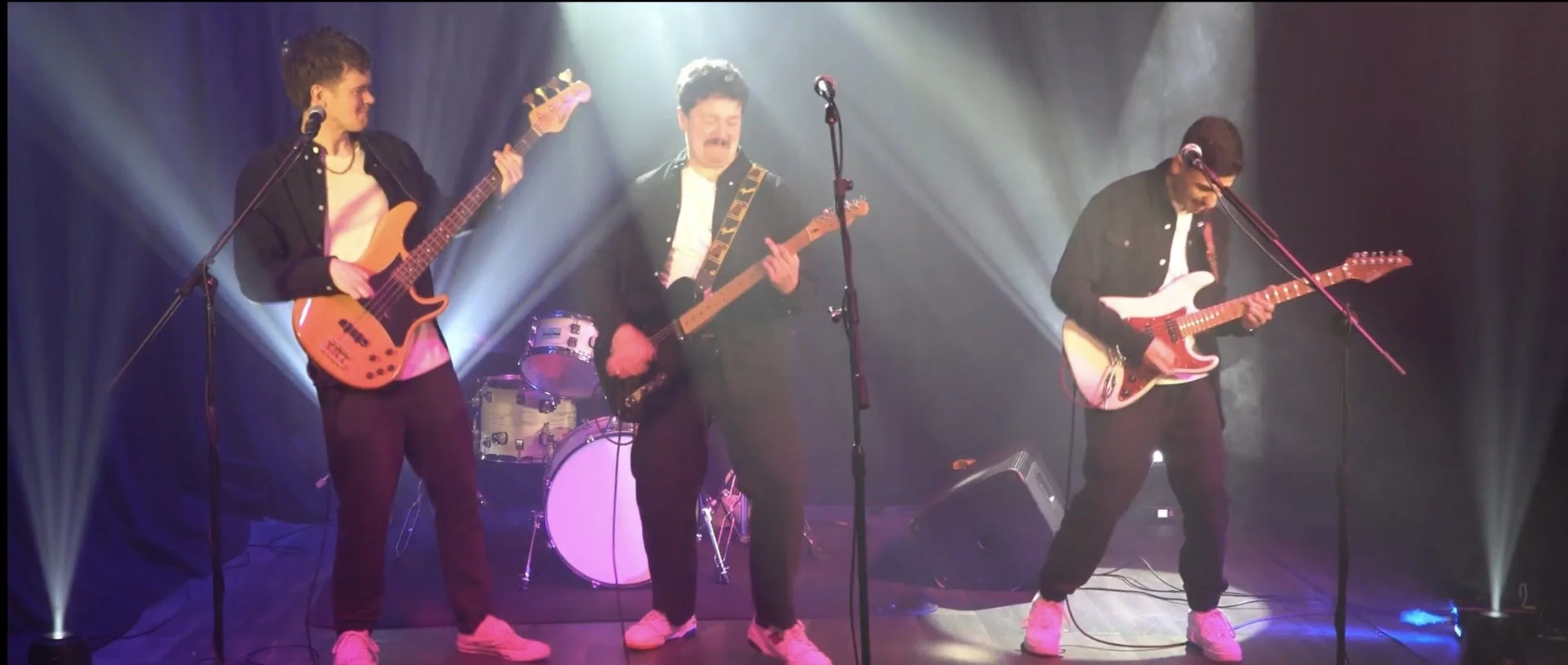 A band of four young men performing on stage with guitars, a drum set in the background, and stage lighting creating a vibrant atmosphere.