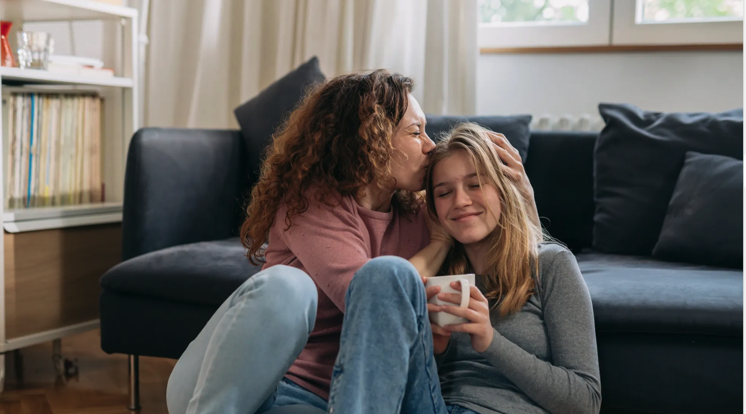A woman with curly hair kisses a girl with long blonde hair on the forehead while they sit on a couch in a living room. The girl holds a mug with both hands, and both appear happy and relaxed.
