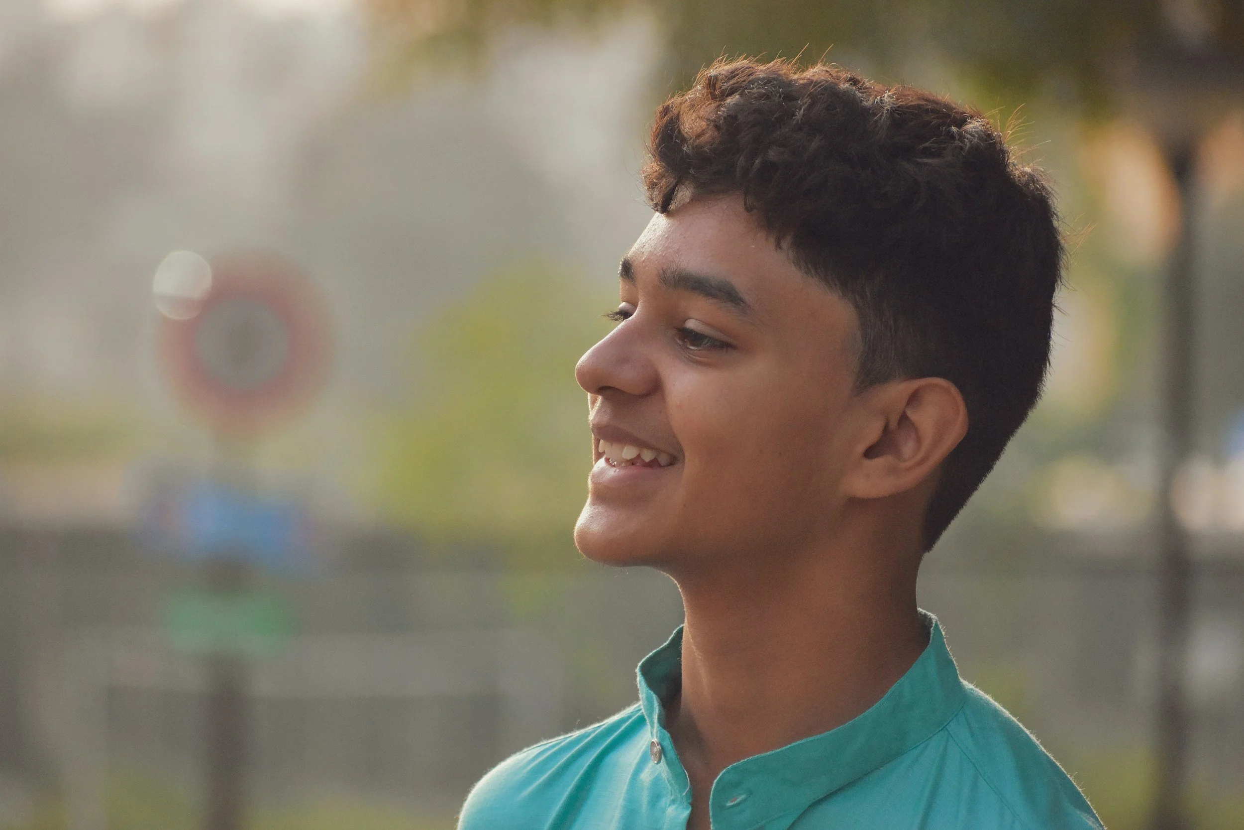 A smiling young man with dark, curly hair and light brown skin, wearing a teal shirt, outdoors in natural light.