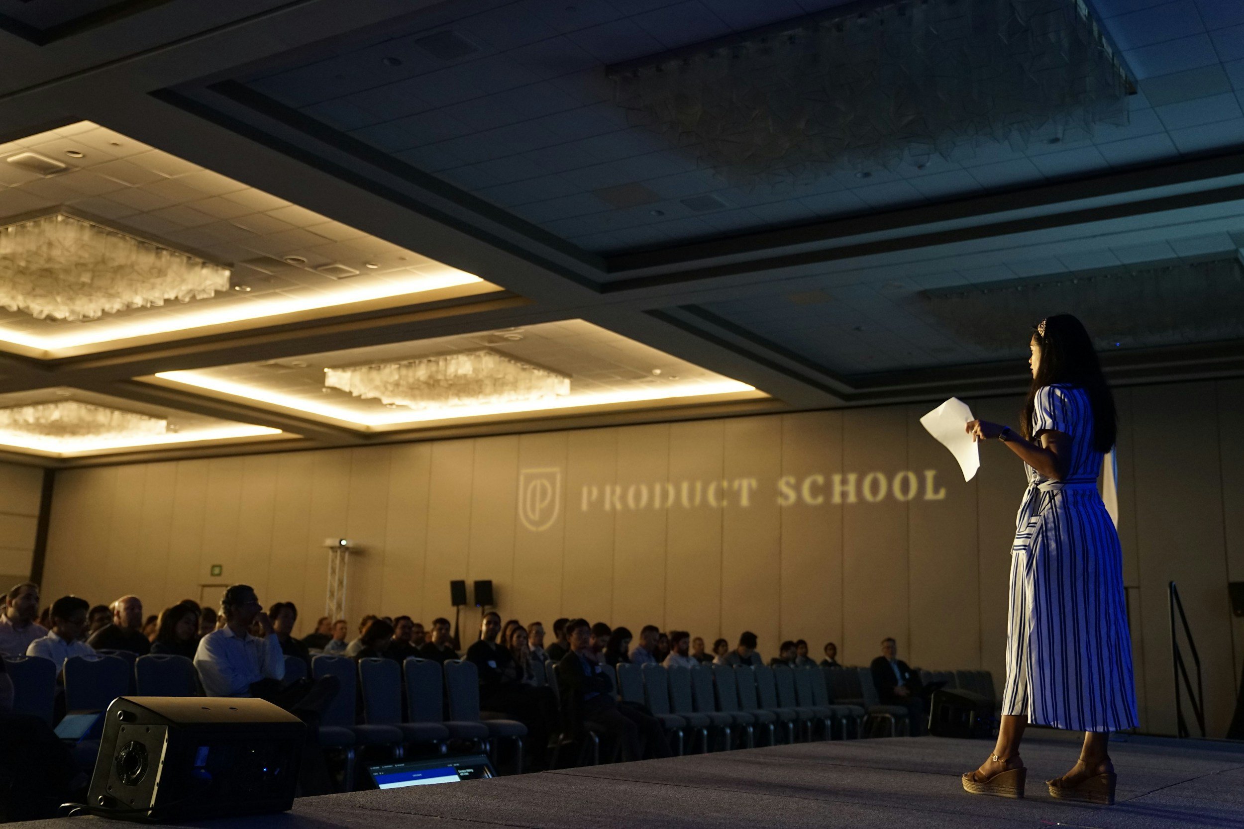A woman standing on stage giving a presentation to an audience seated in a conference room with decorated ceilings and a sign that reads 'Product School' in the background.