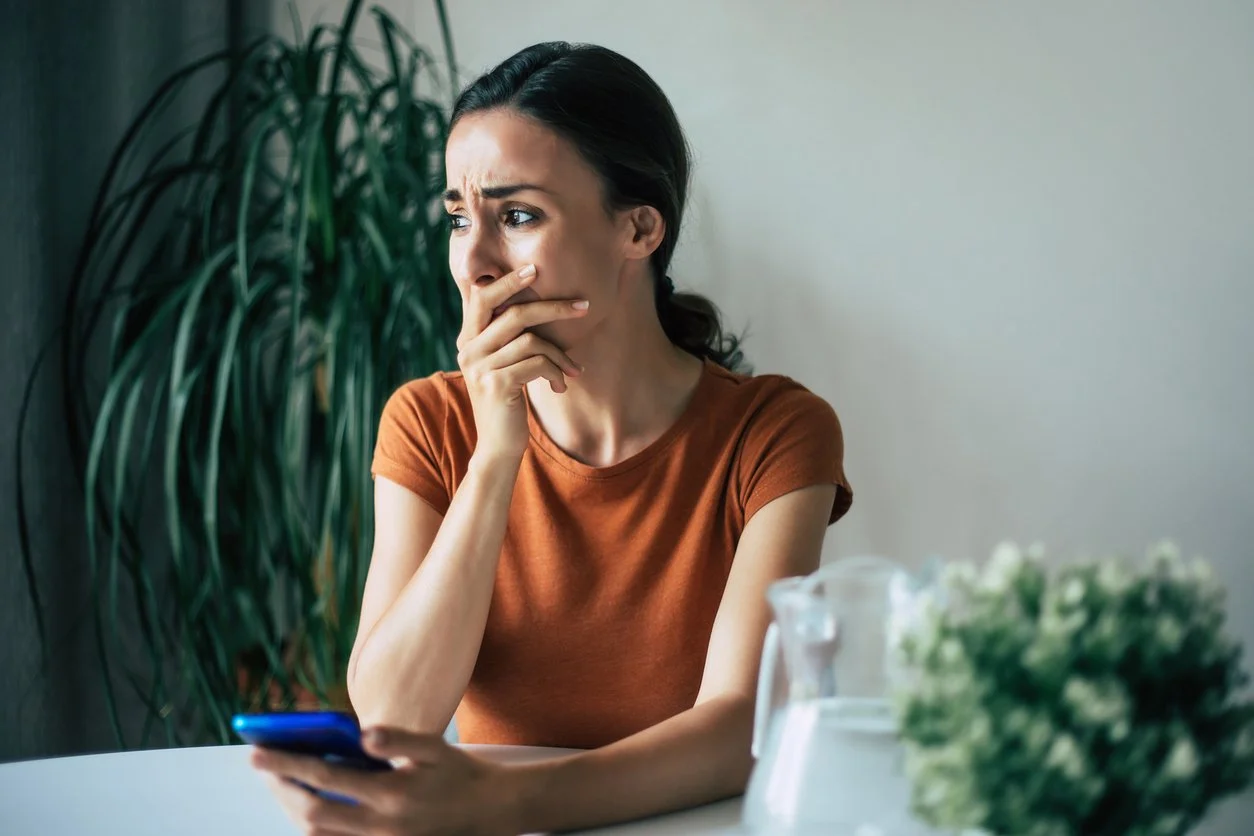 A woman sitting at a table looking at her phone with a worried or confused expression, holding her hand over her mouth. There are a large green plant in the background and some blurred objects, possibly a flower or decoration, on the table.