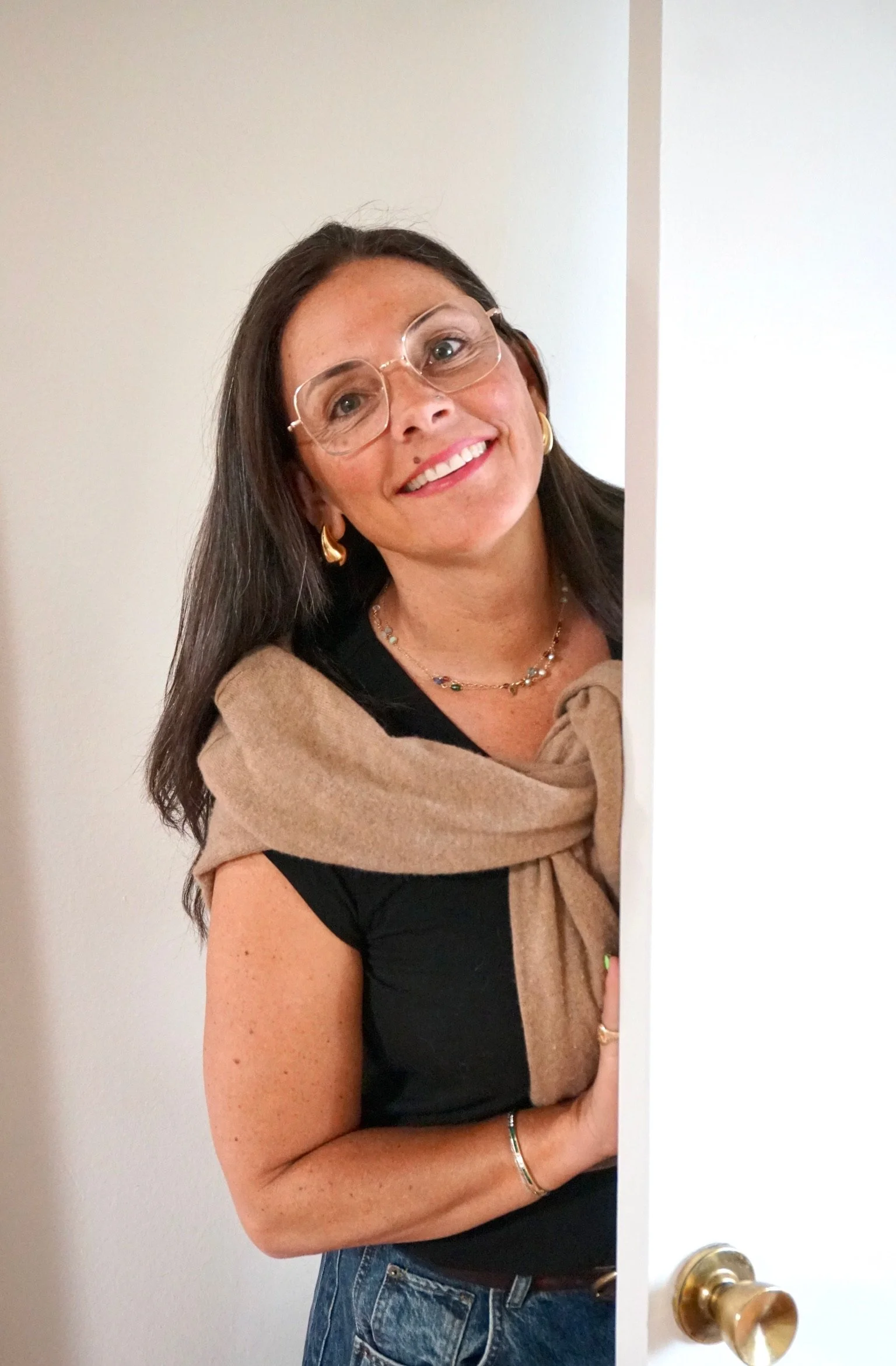 A woman with long dark hair, wearing glasses, gold earrings, a necklace, a bracelet, a black shirt, and jeans, smiling while peeking from behind a door.