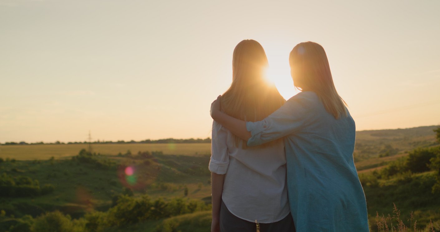 Two women , a mother and a daughter, with light hair standing outdoors during sunset, embracing and looking at the horizon over a rural landscape with fields and trees.