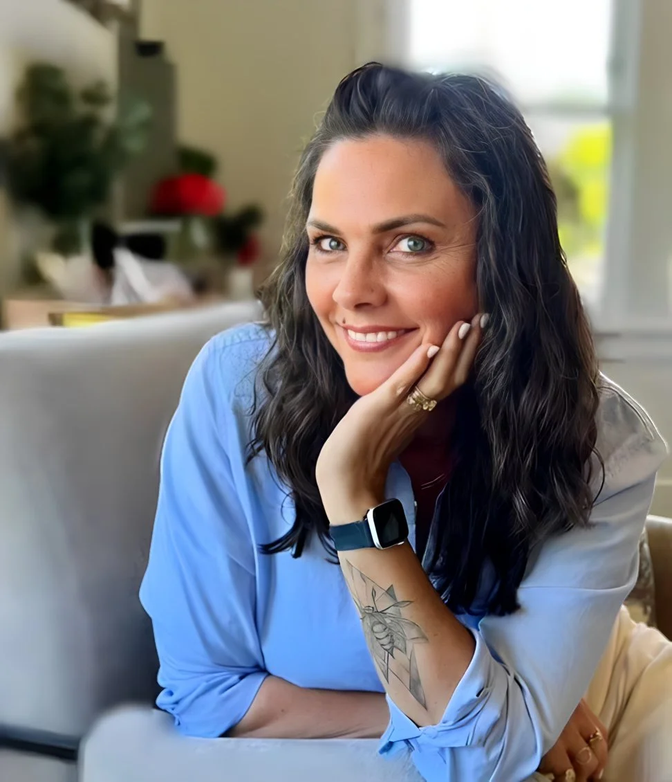 A woman with dark, wavy hair and blue eyes, smiling at the camera, resting her chin on her hand, wearing a light blue shirt with the sleeves rolled up, a smartwatch, and a tattoo on her forearm, seated in a cozy, well-lit room.