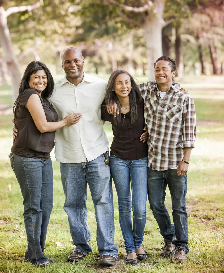 A diverse group of four people, two women and two men, standing outdoors in a park with trees. They are smiling and embracing each other, looking happy and enjoying the sunshine.