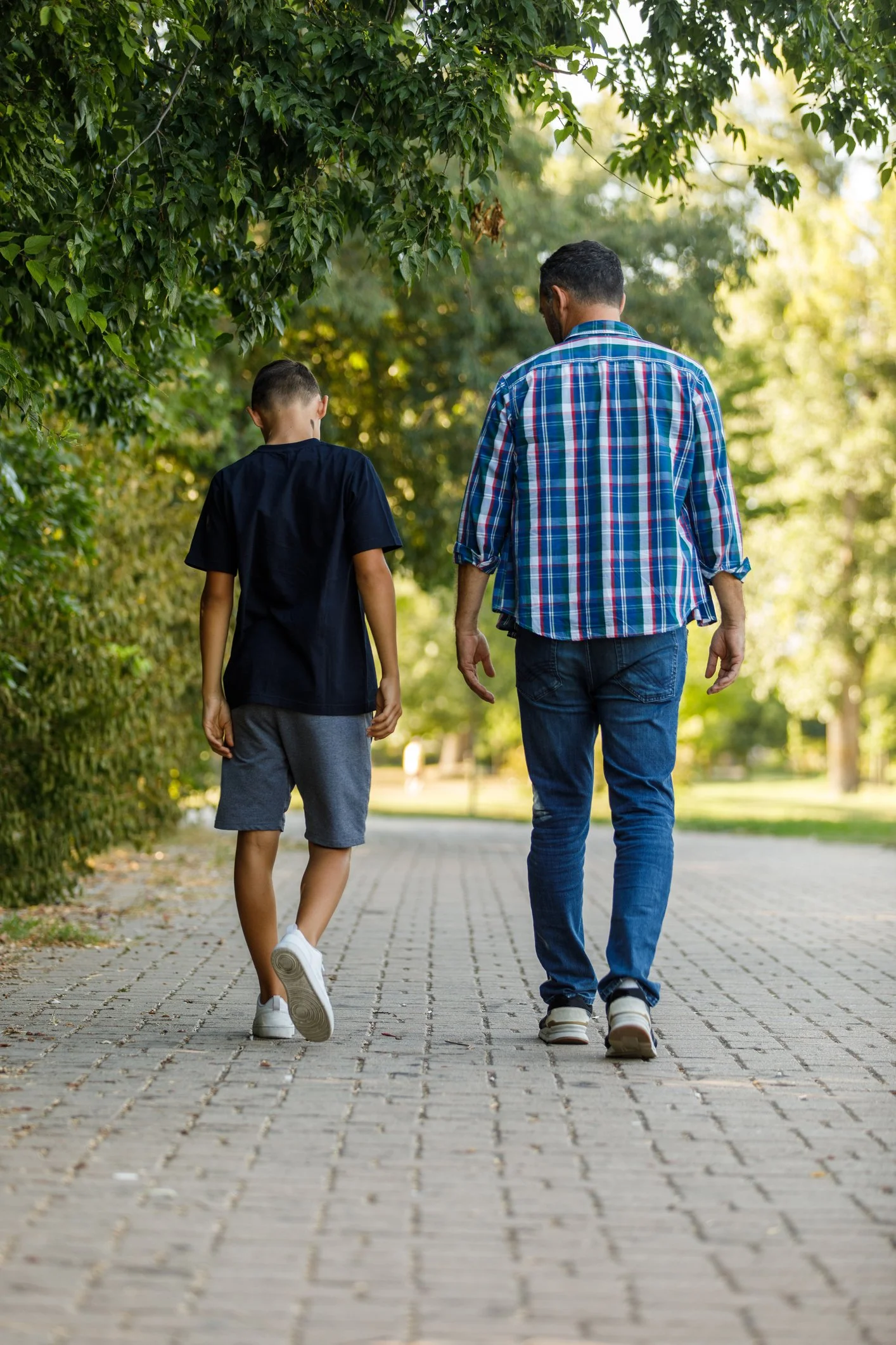 A man and a boy walking together on a paved path surrounded by lush greenery in a park.