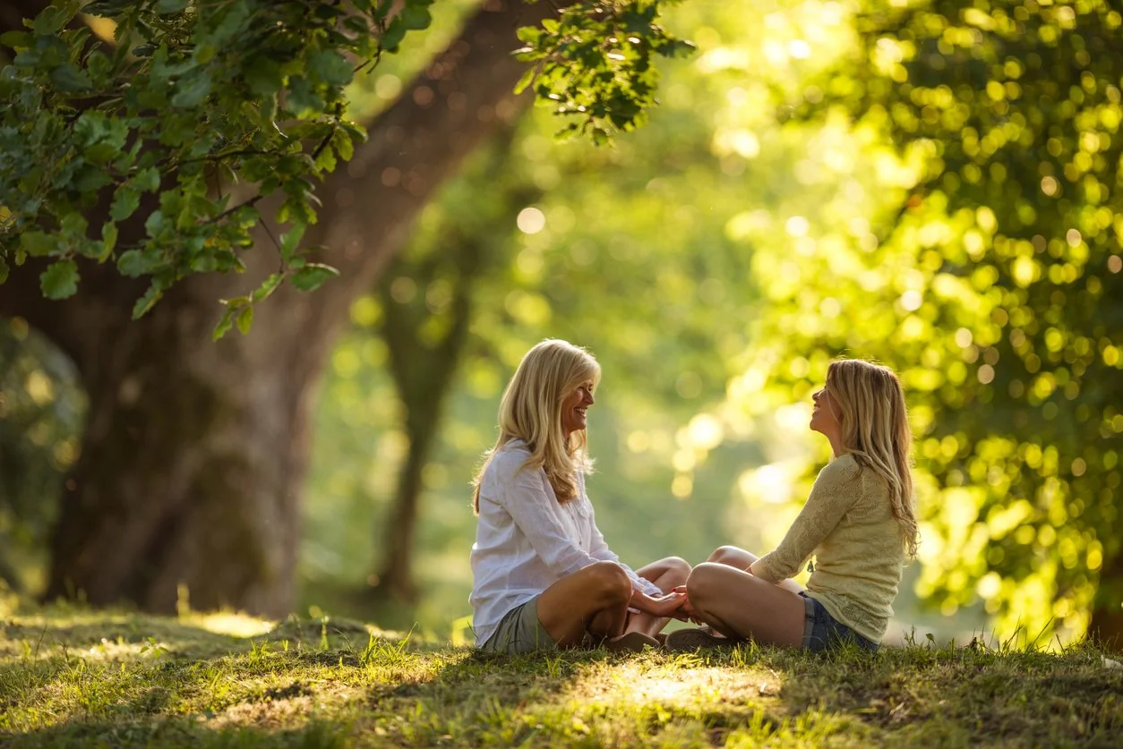 A mother and daughter, sitting cross-legged on the grass in a park with green trees, enjoying a sunny day and sharing a happy moment.
