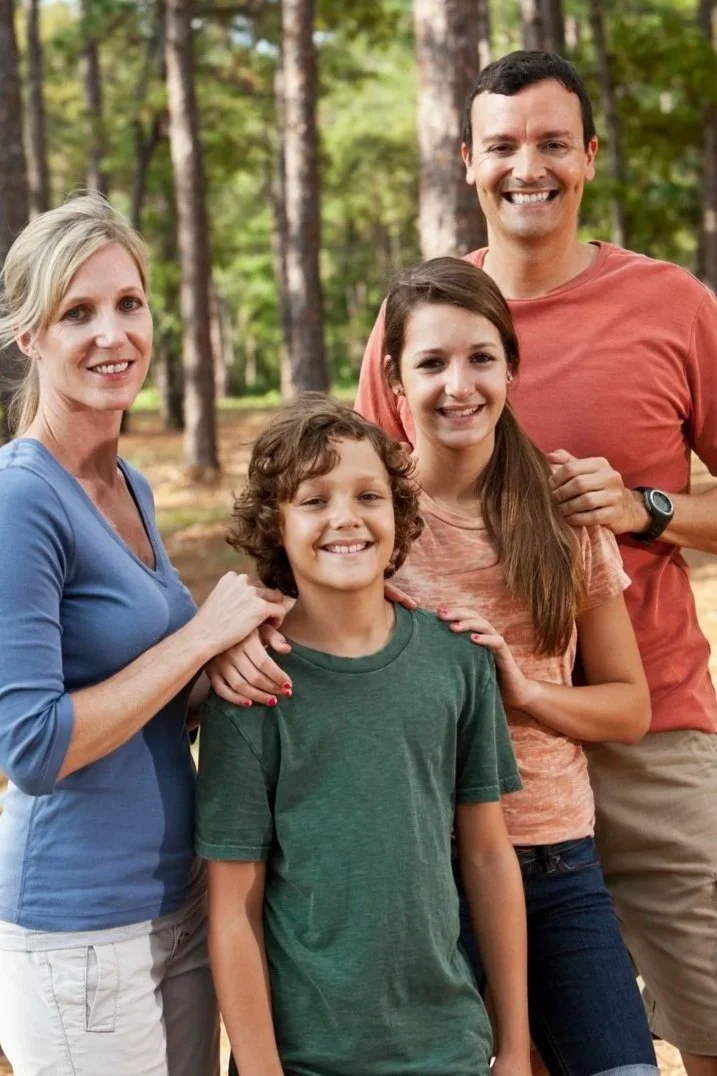 A family of four posing outdoors in a wooded area, smiling at the camera.
