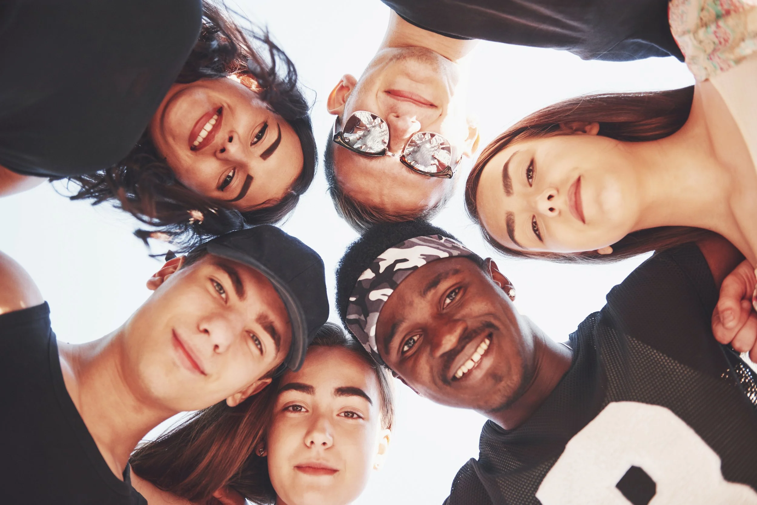 A diverse group of people standing in a circle, looking down into the camera, smiling and showing a sense of unity and friendship.