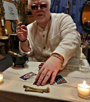 Person wearing sunglasses and a beige shirt, sitting at a table with tarot cards, a candle, and a small pot, in a dimly lit setting.