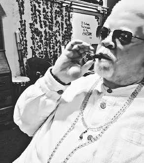 Man with sunglasses and a white shirt, smoking a cigar, wearing a chain around his neck, in an indoor setting.