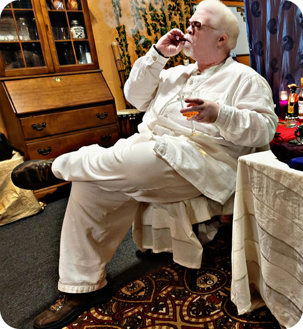 A man with white hair and glasses sitting comfortably in a dining room, drinking from a small glass of what appears to be alcohol, with a relaxed pose and a thoughtful expression.