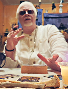 Man with white hair and sunglasses sitting at a table with tarot cards, a bone object, and a drink, gesturing with her hand in a room decorated with hanging items and shelves.