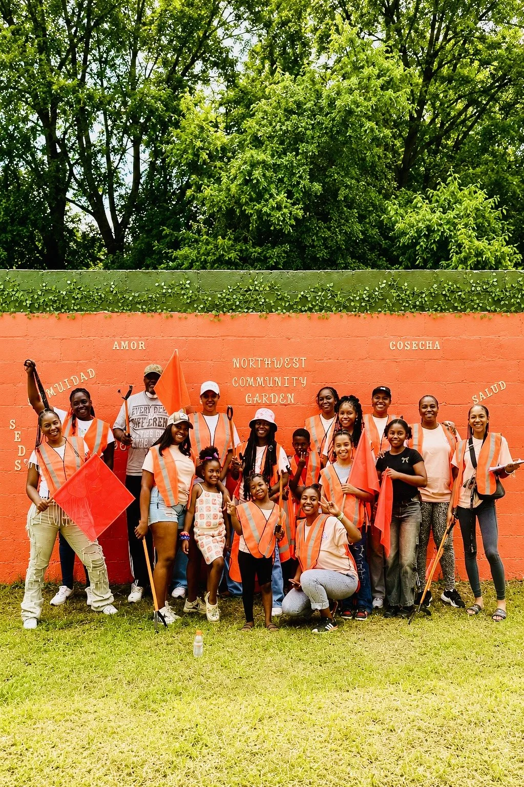 Group of diverse people, mostly young girls, wearing orange safety vests and holding red flags, standing in front of a brightly painted orange wall with various words in Spanish and English, including 'Amor', 'Cosecha', 'Salud', and 'NORTHWEST COMMUNITY GARDEN', with green trees behind the wall.