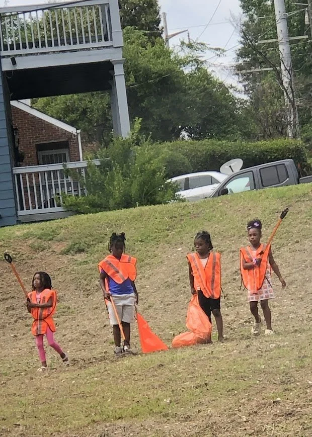 Four young girls wearing orange safety vests standing on a grassy hill, holding litter pickers and trash bags, participating in a community cleanup activity.
