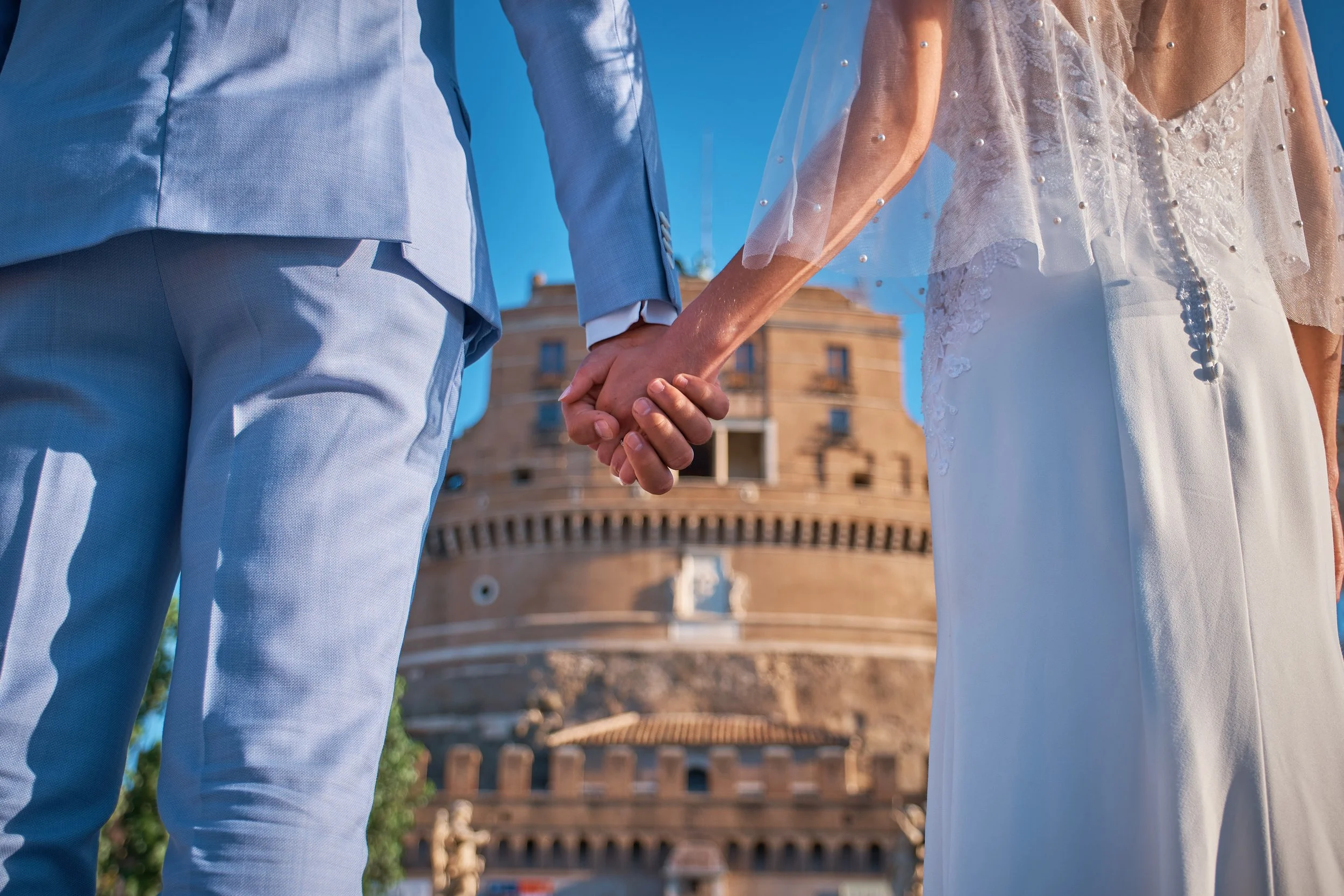 A couple holding hands in front of the Colosseum in Rome, Italy, with the sky visible above.