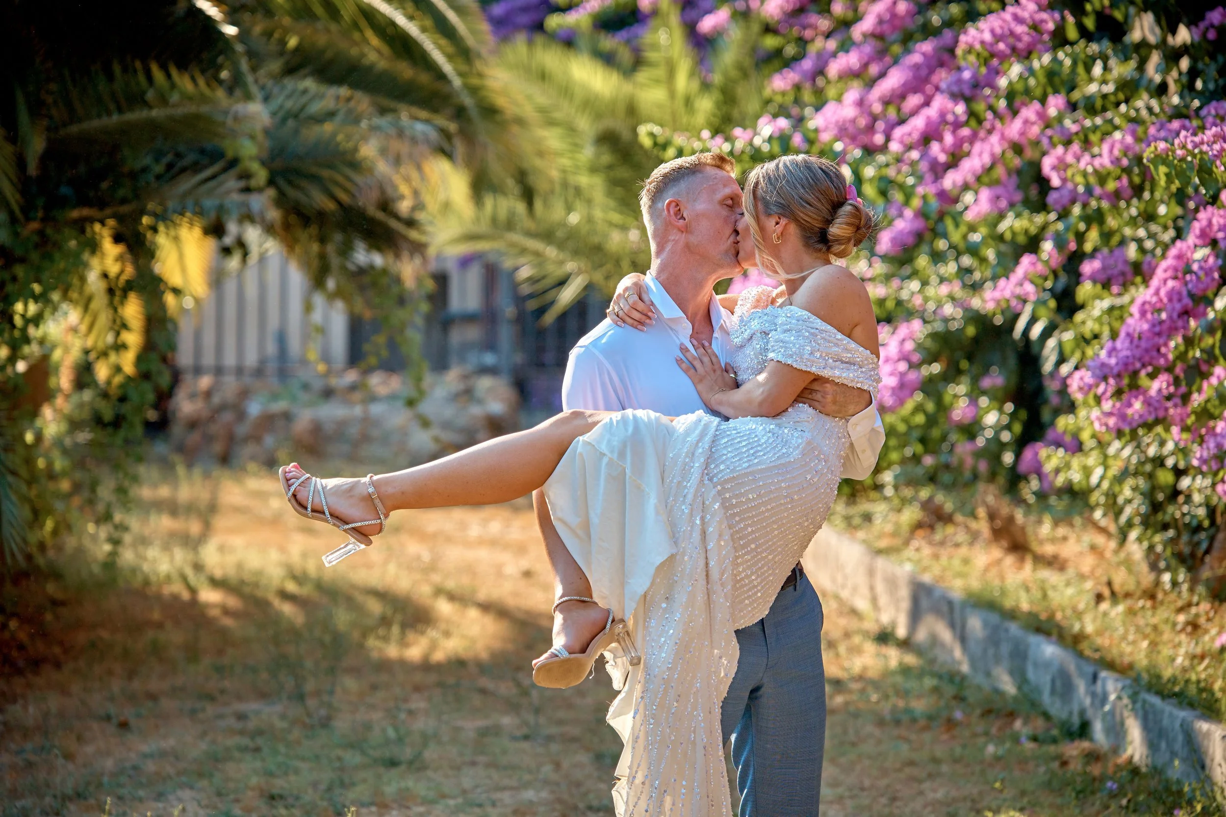 A man lifting and kissing a woman in a white dress outdoors, surrounded by pink and purple flowering bushes and palm trees.