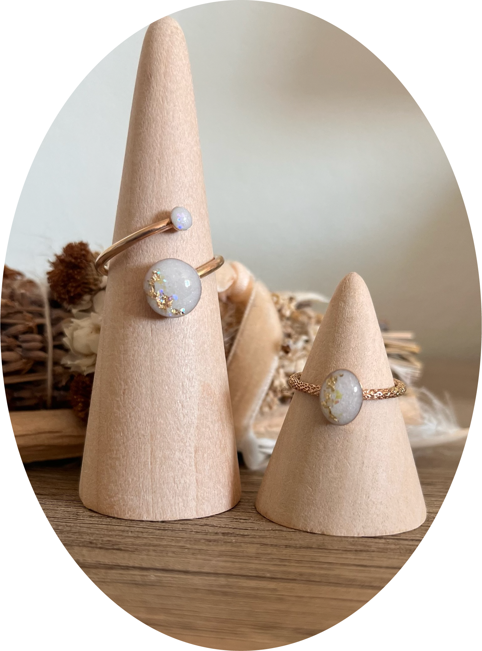 Three beige ring displays on cone-shaped jewelry holders, with a background of dried flowers and a basket.