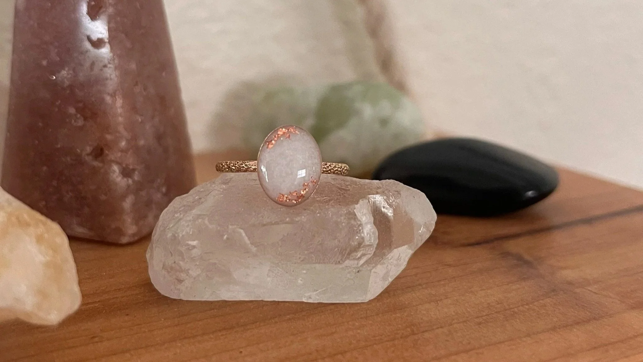 A close-up of a gold ring with a large, oval, transparent gemstone adorned with small pinkish granules, placed on a clear quartz crystal. In the background, there's a brownish stone, a black object, and green leaves, all on a wooden surface.