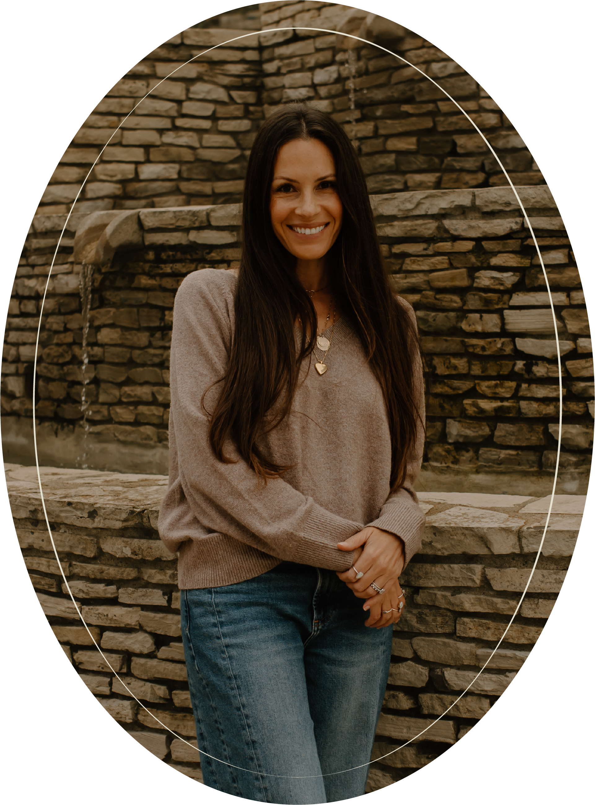 A woman with long dark hair smiling, standing in front of a stone water fountain, wearing a beige sweater and blue jeans.