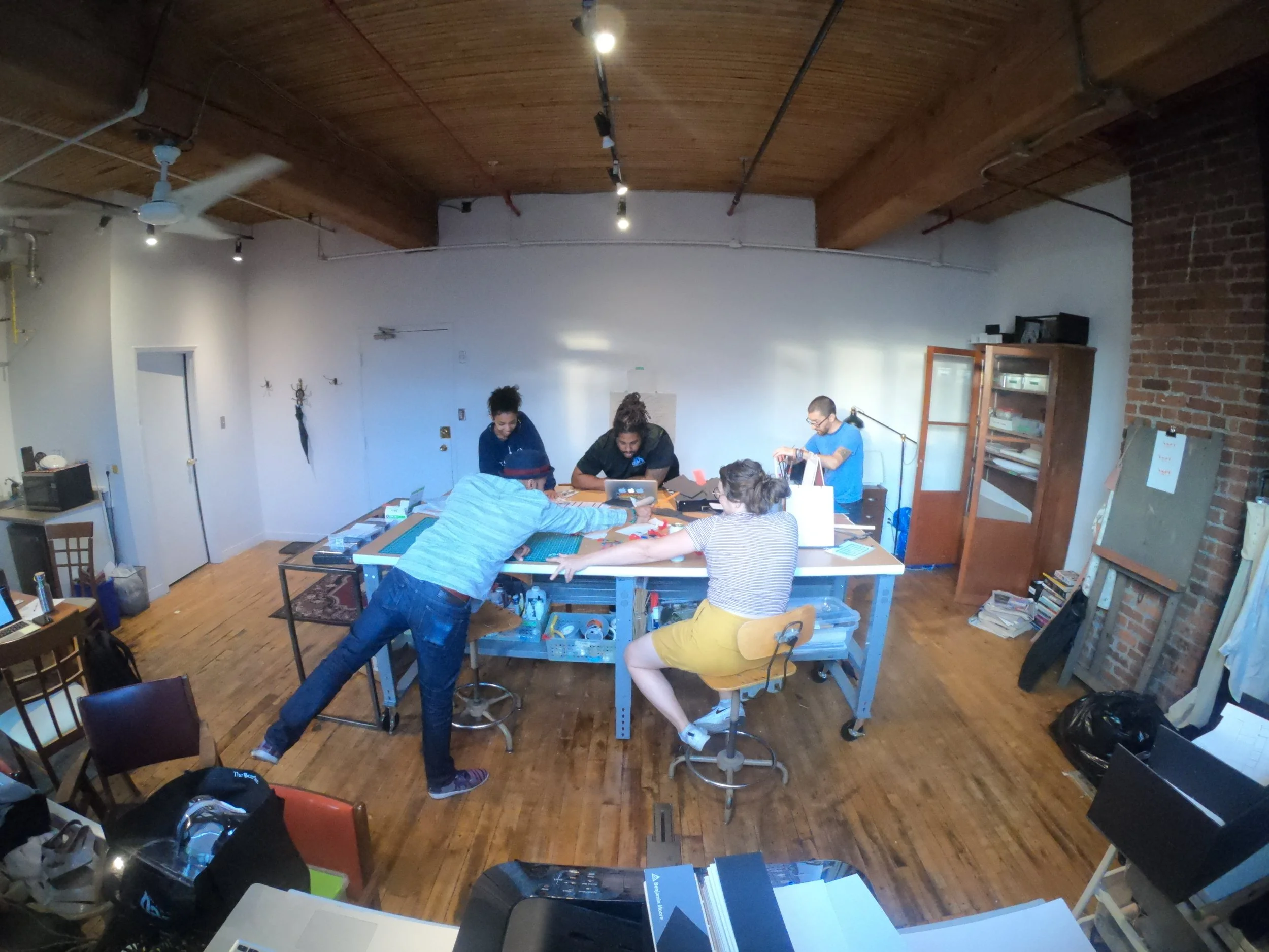 A group of six people gathered around a table in a room with wooden floors and a wooden ceiling. They appear to be working on a project with various tools and materials on the table. The room has an industrial style with brick and white walls, and some shelves and furniture. One person is standing while the others are seated, some leaning over the table.